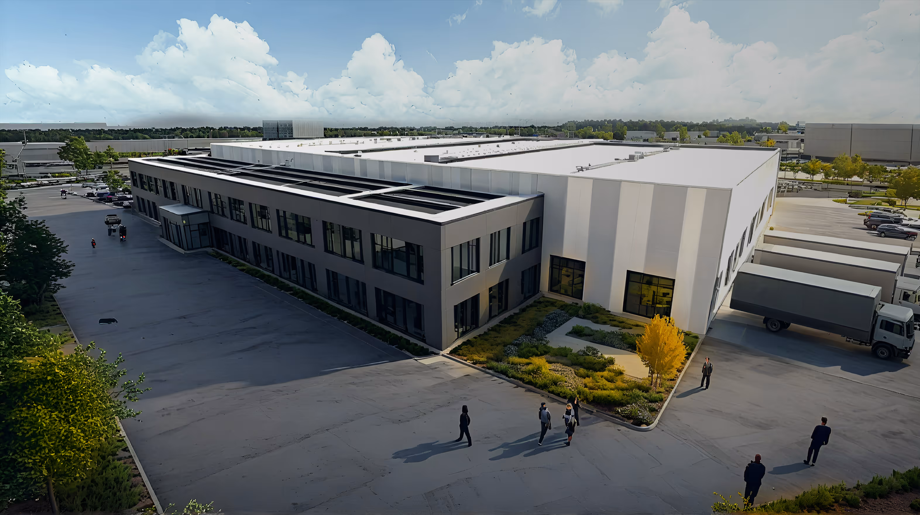 A large modern industrial building with a gray office section and white warehouse area, surrounded by parking lots, trucks, and a few people walking nearby under a partly cloudy sky.