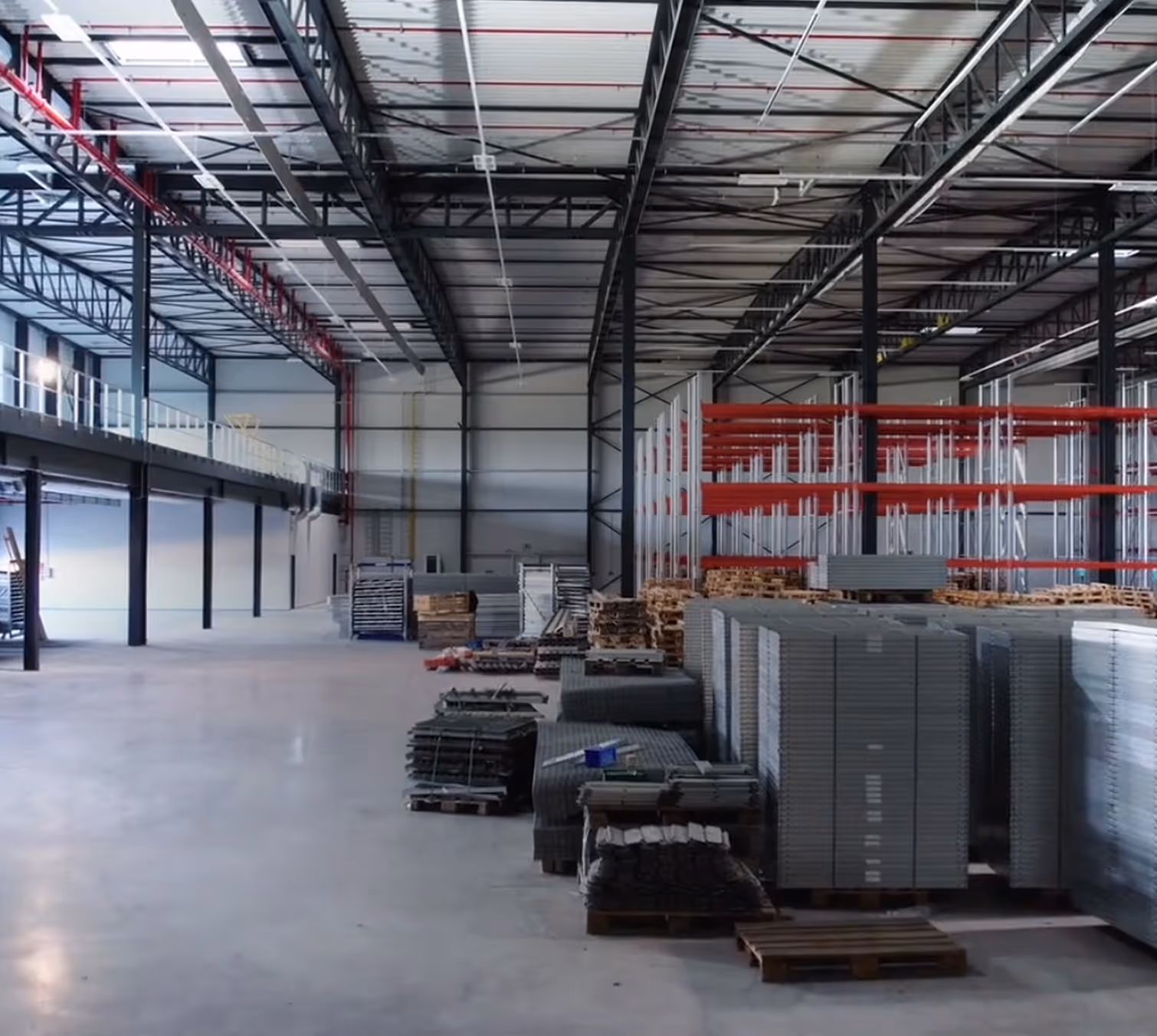 Spacious warehouse with stacked pallets and metal shelving racks under a steel beam ceiling.