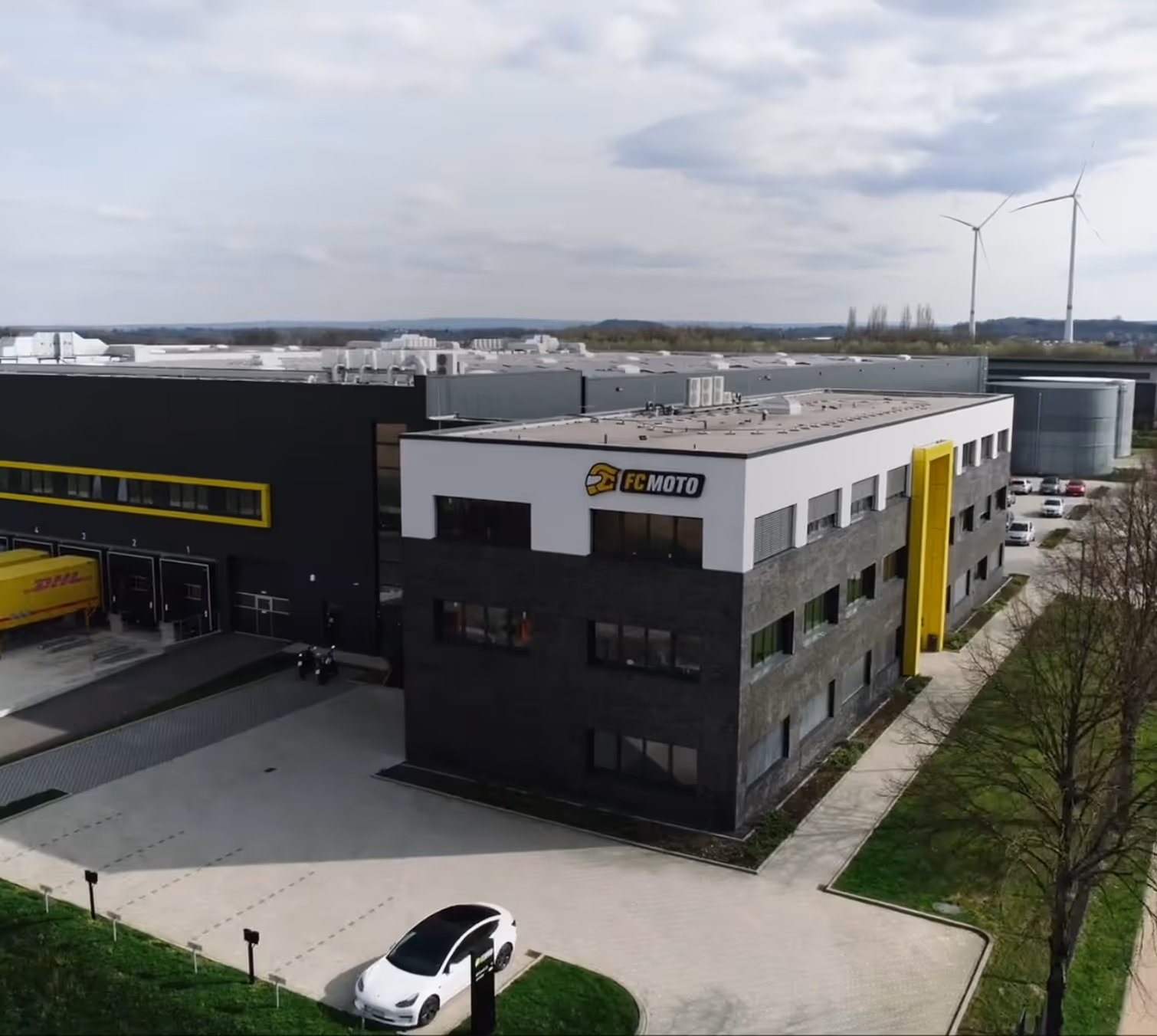 Modern FC-Moto building with black and white facade and yellow accents, parked white car in foreground, and wind turbines in background.