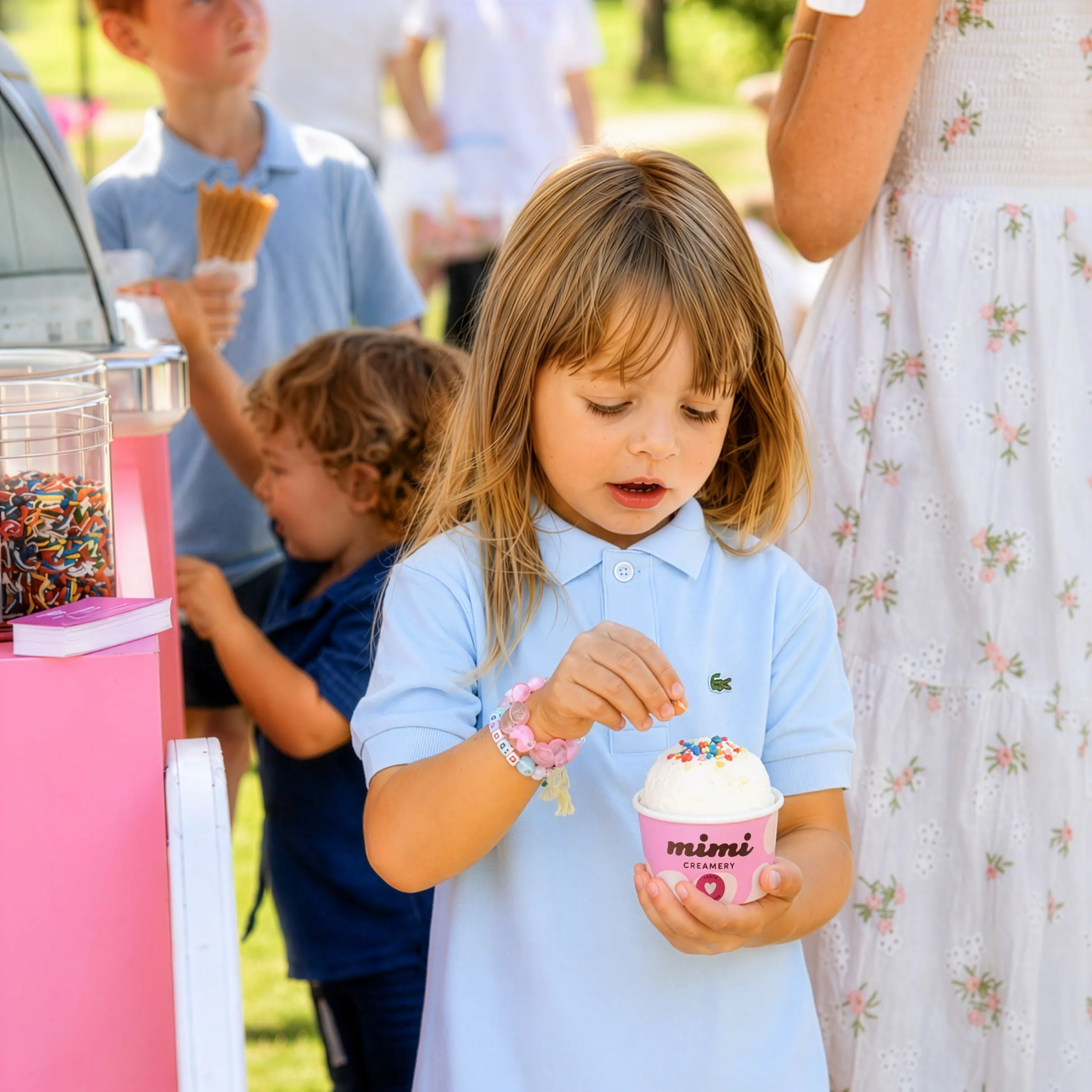 Child in blue shirt adding sprinkles to ice cream in a pink Mimi Creamery cup outdoors.
