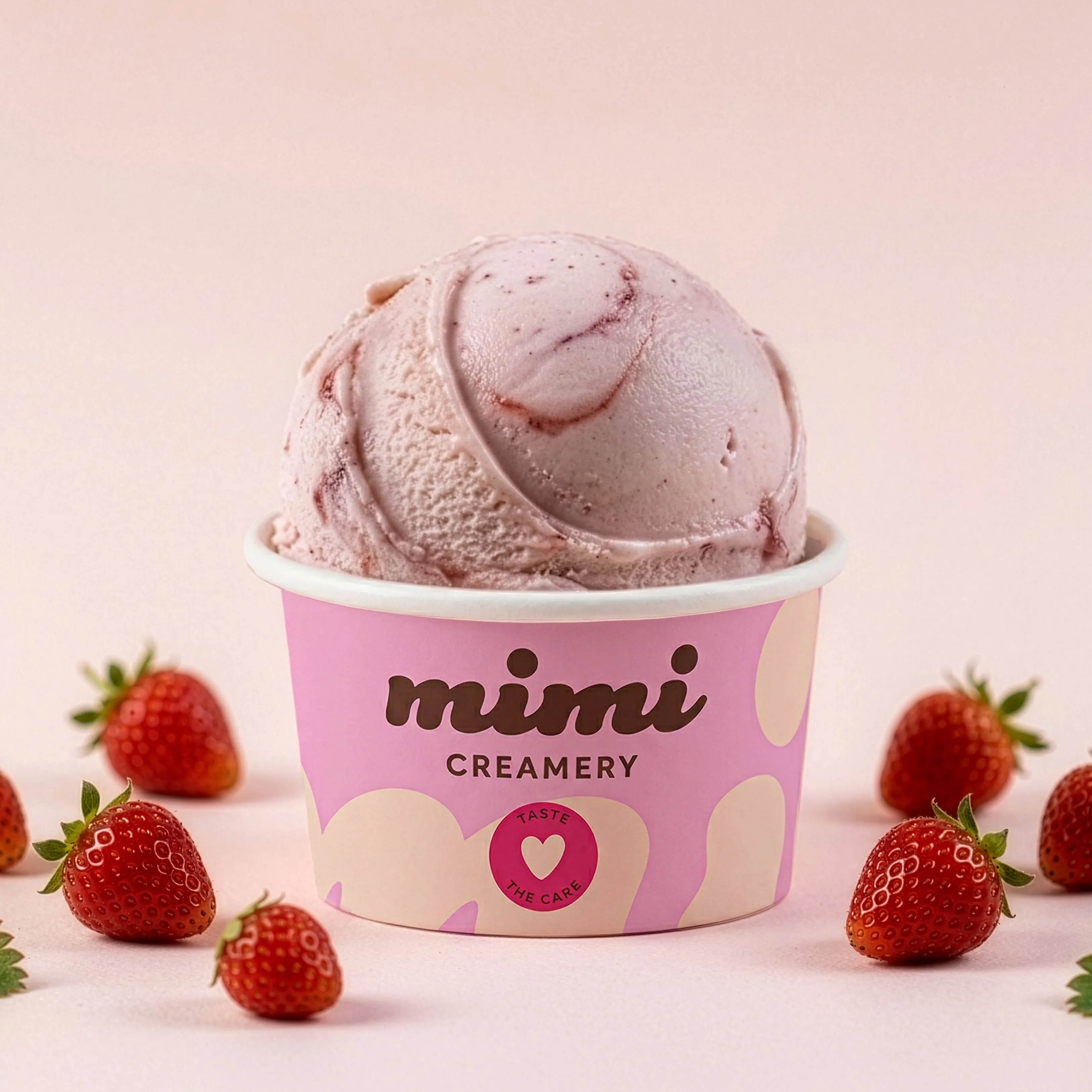Pink strawberry ice cream scoop in a pink Mimi Creamery cup surrounded by fresh strawberries.