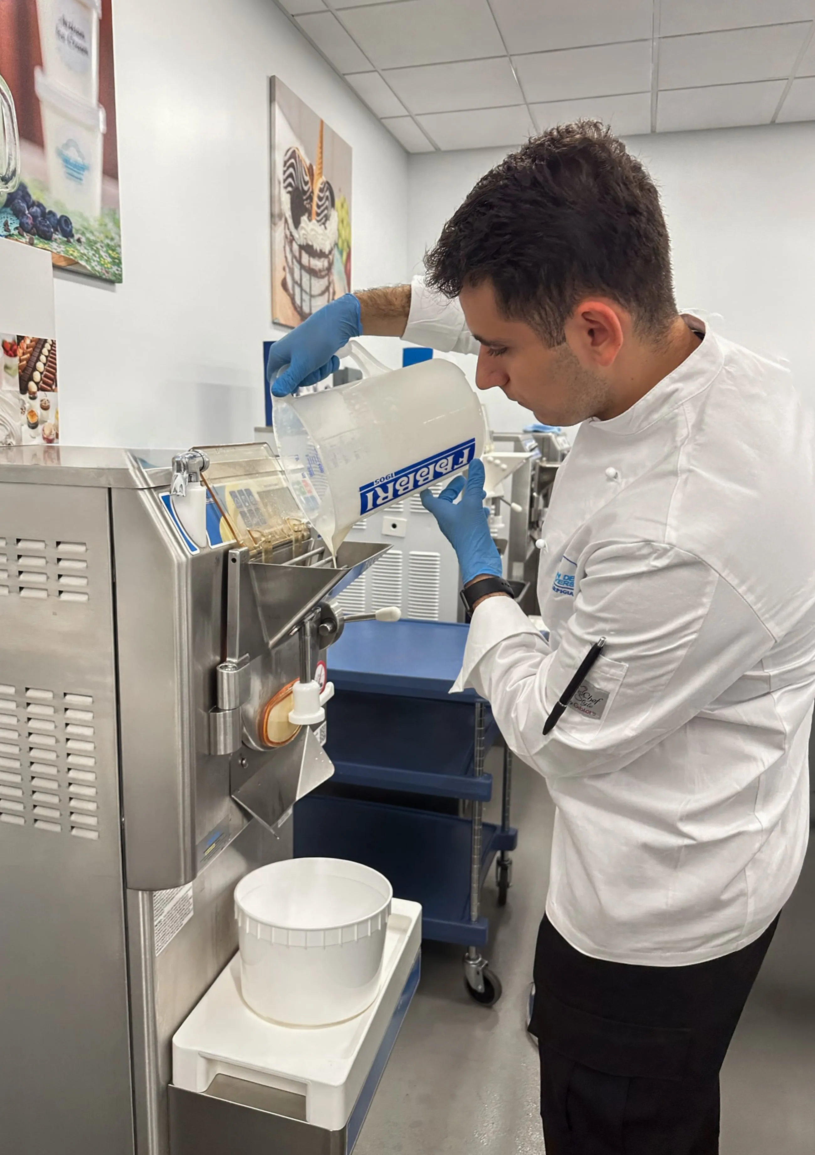 Man in white chef coat pours liquid from a measuring cup into stainless steel machine in kitchen.