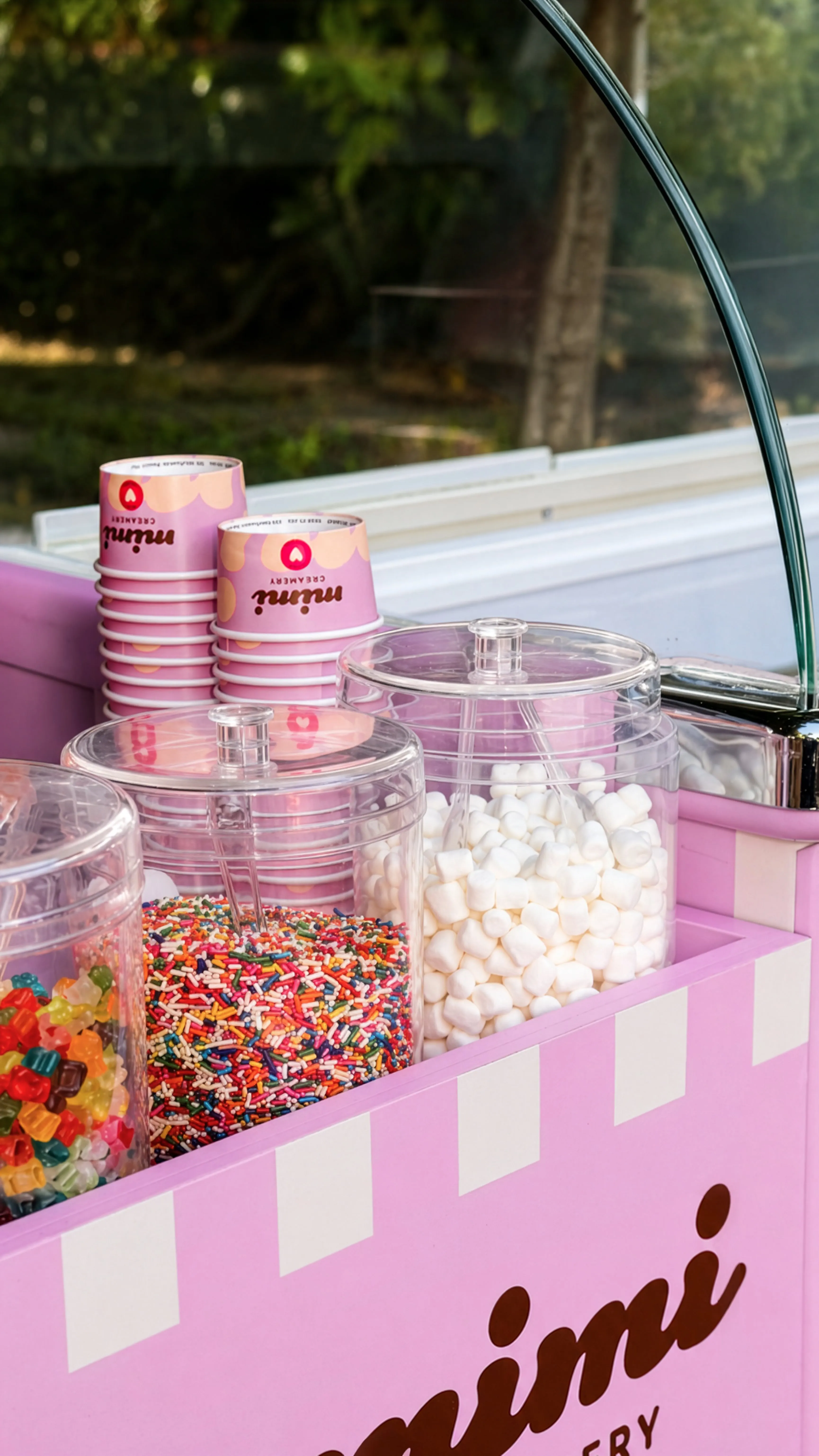 Pink dessert topping station with containers of marshmallows, sprinkles, gummy bears, and pink ice cream cups.