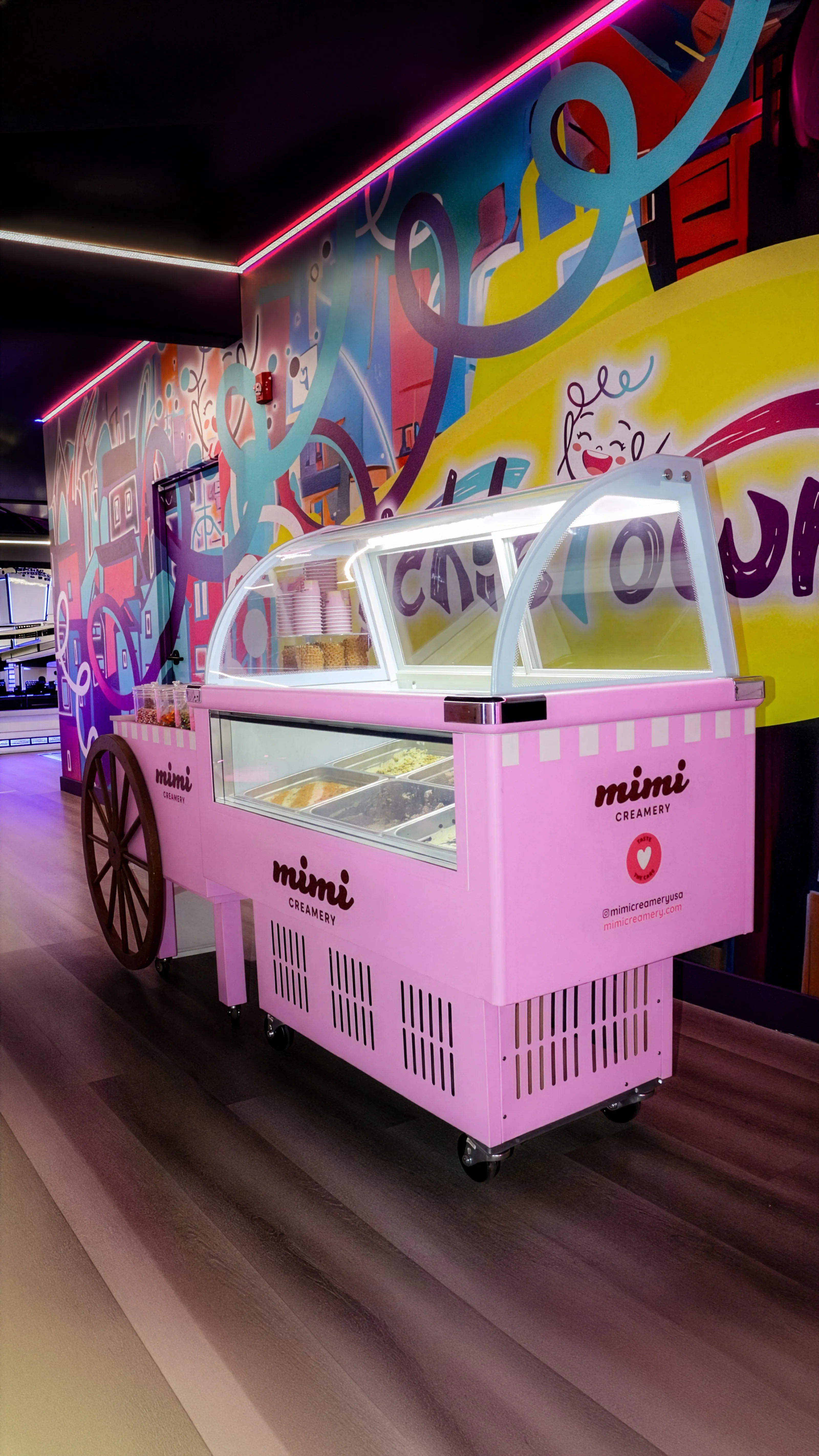 Pink ice cream cart with Mimi Creamery logo in a colorful room with mural wall art and neon lights.