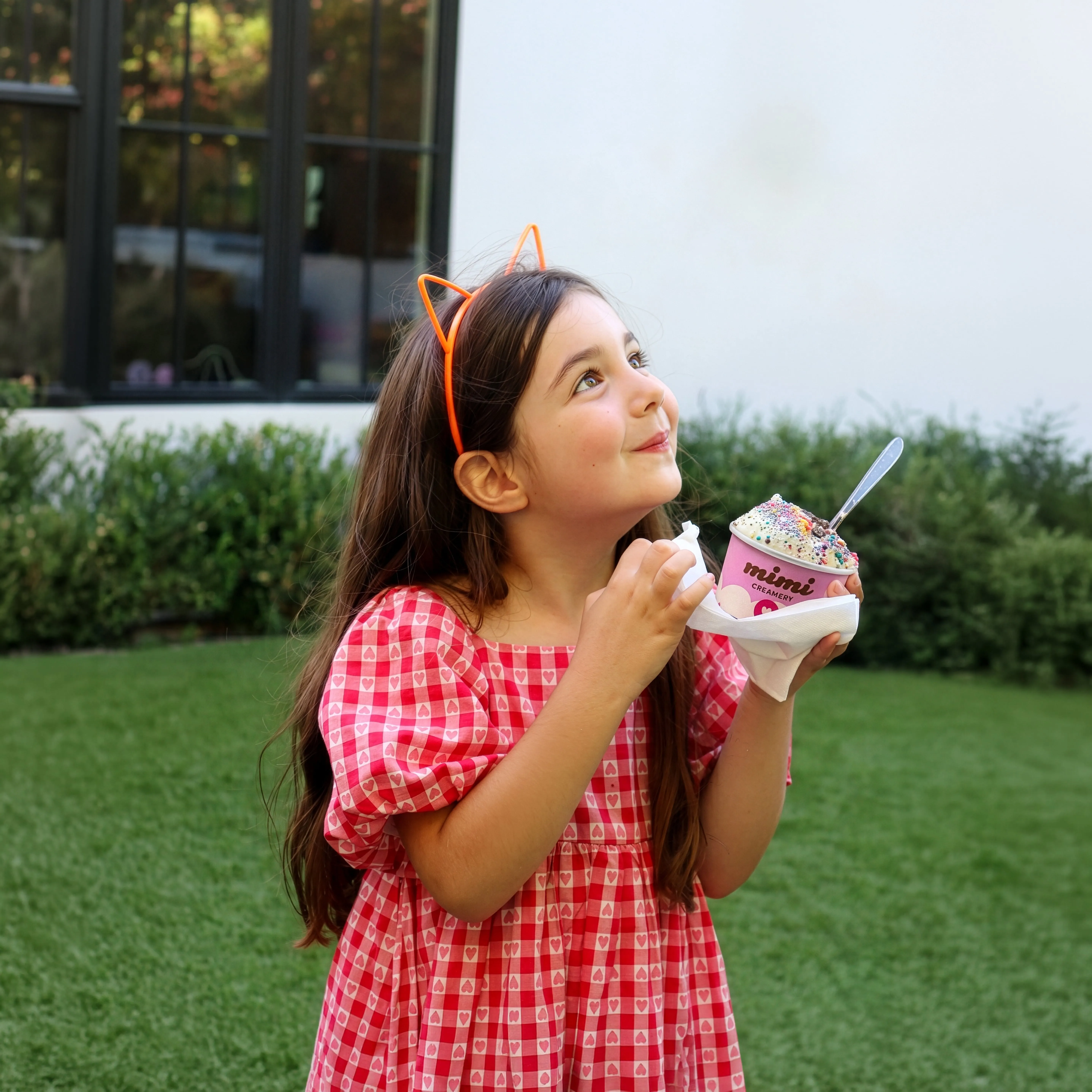 Young girl with orange cat ear headband eating colorful sprinkled ice cream outside on grass.