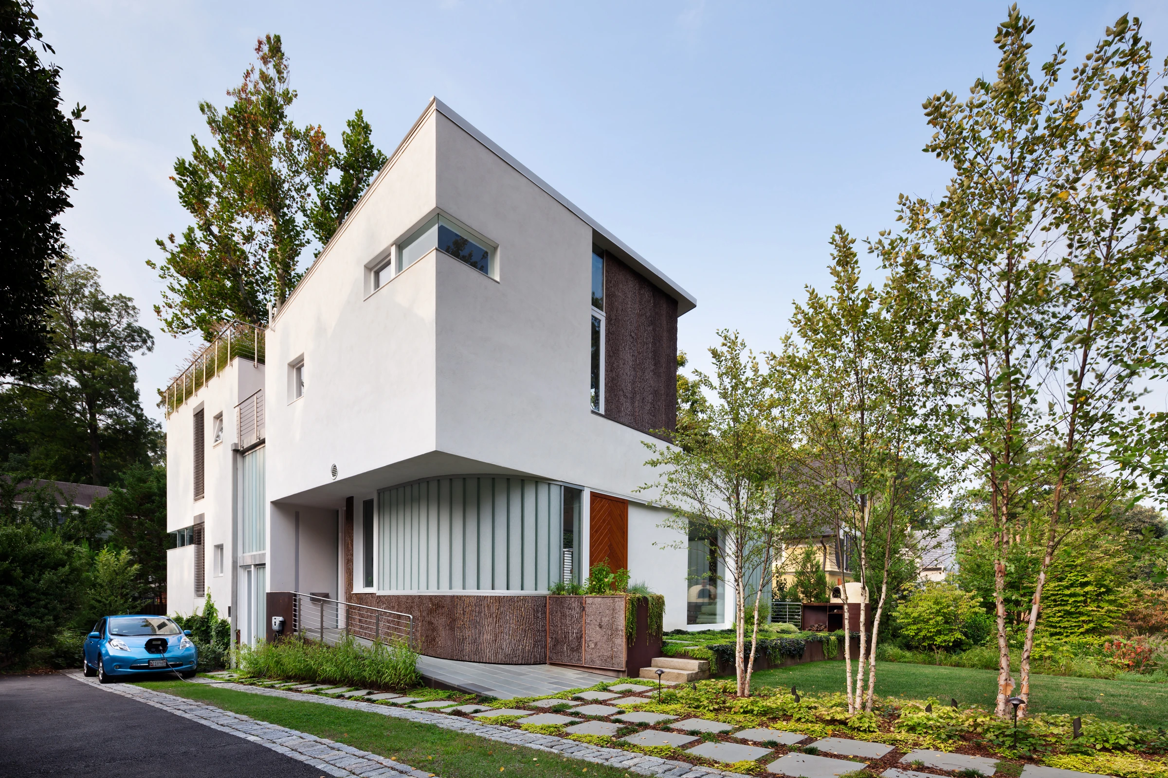 3/4 view of the house and driveway, showing the electric car charger and white stucco house with some bark cladding and channel glass.