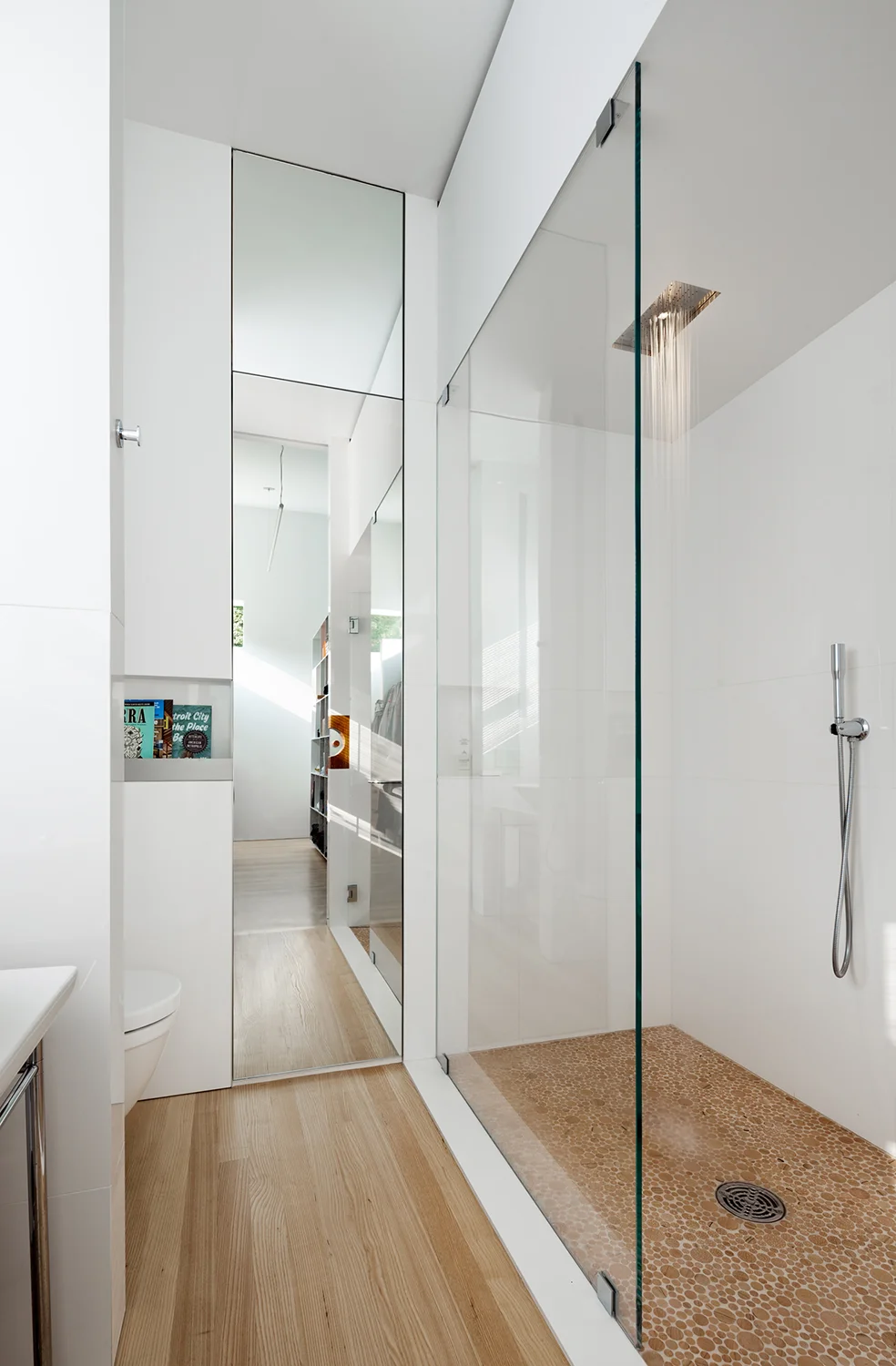 The sleek primary bathroom vanity with round pivoting mirror in front of a window, floor to ceiling mirrored medicine cabinet, and a rain shower.