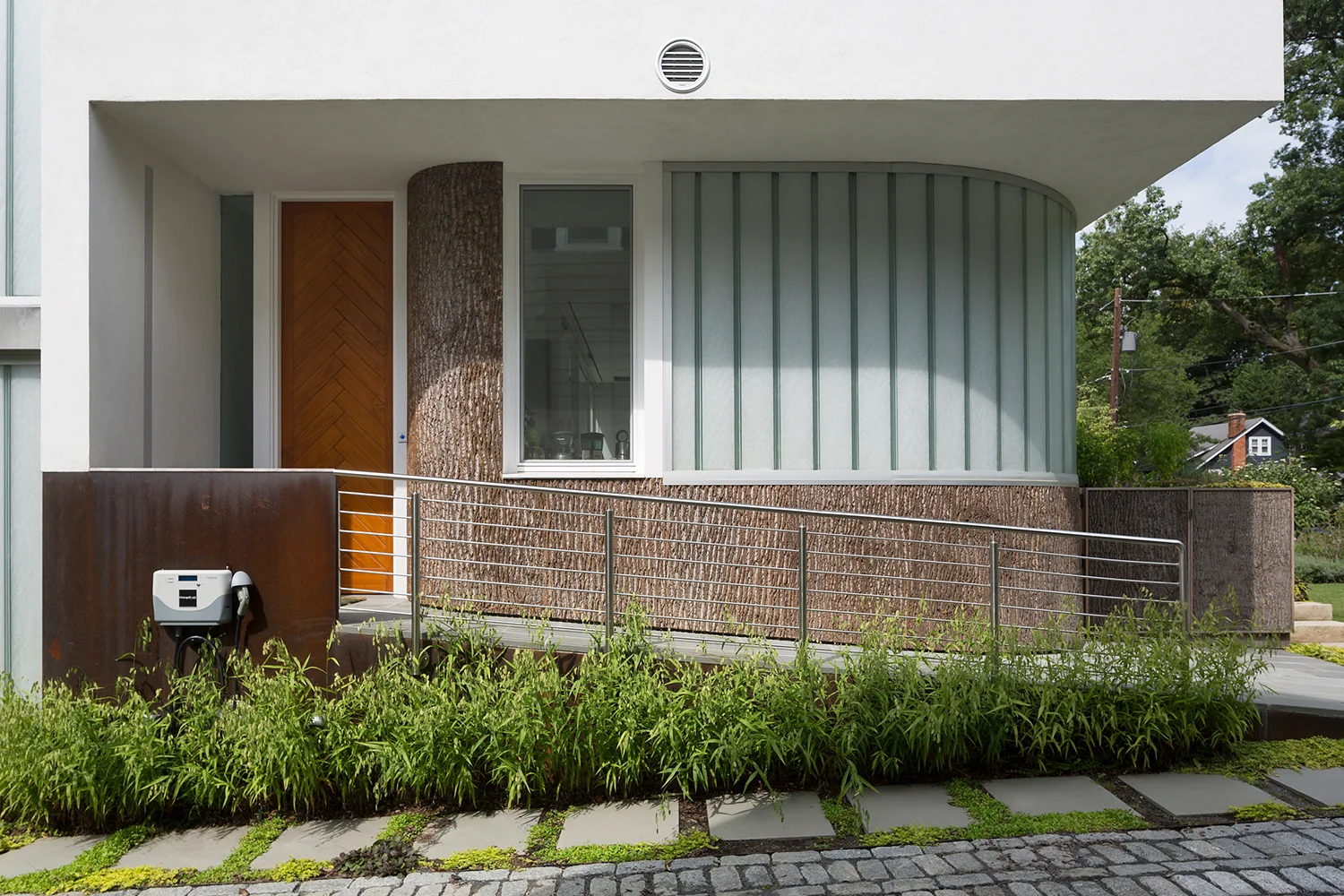 Entry ramp with railing next to the kitchen wall, which is clad in bark with an operable window and curved channel glass.