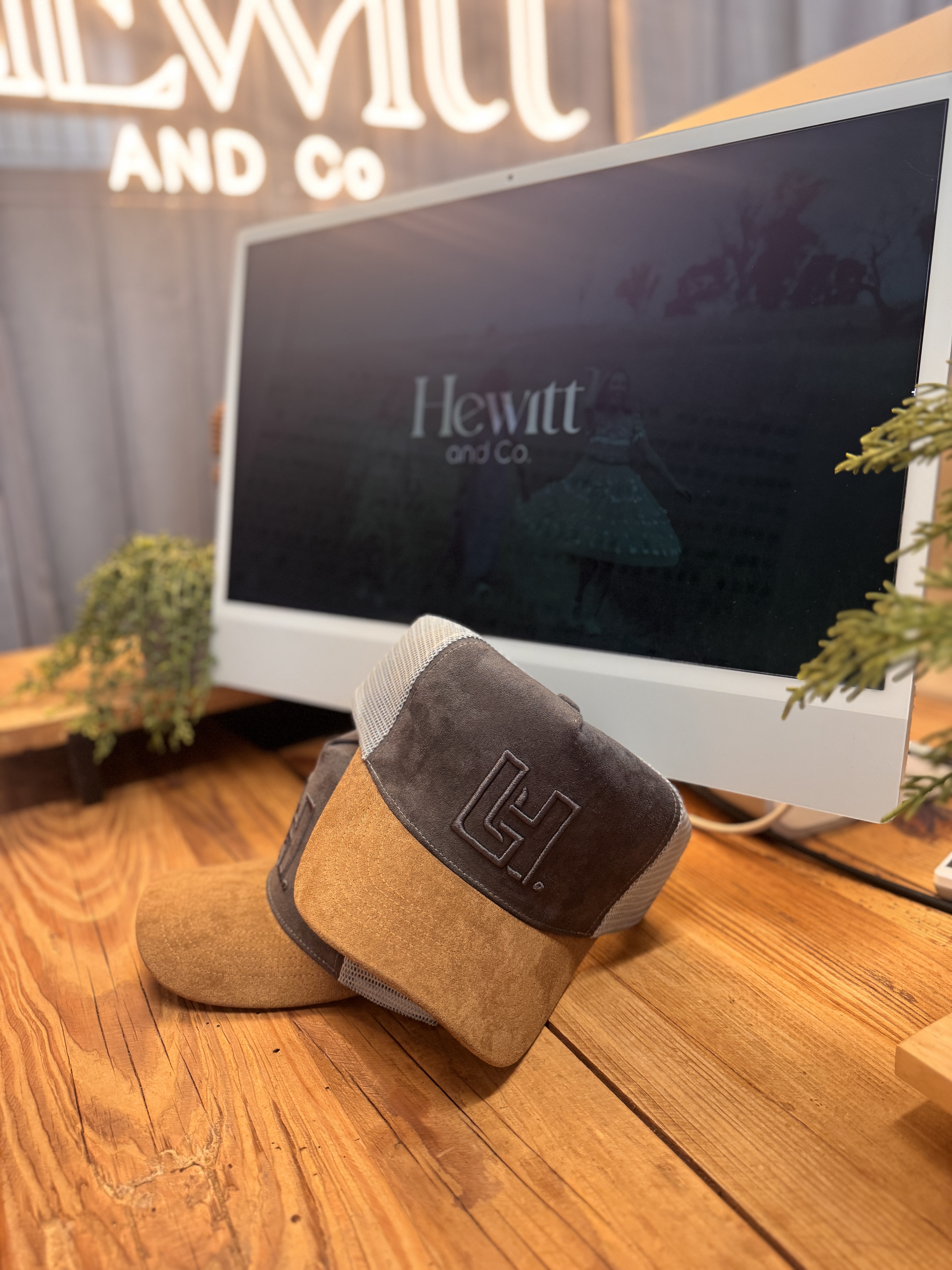 Two brown and beige caps with embroidered logos resting on a wooden desk in front of a computer screen showing 'Hewitt and Co.'