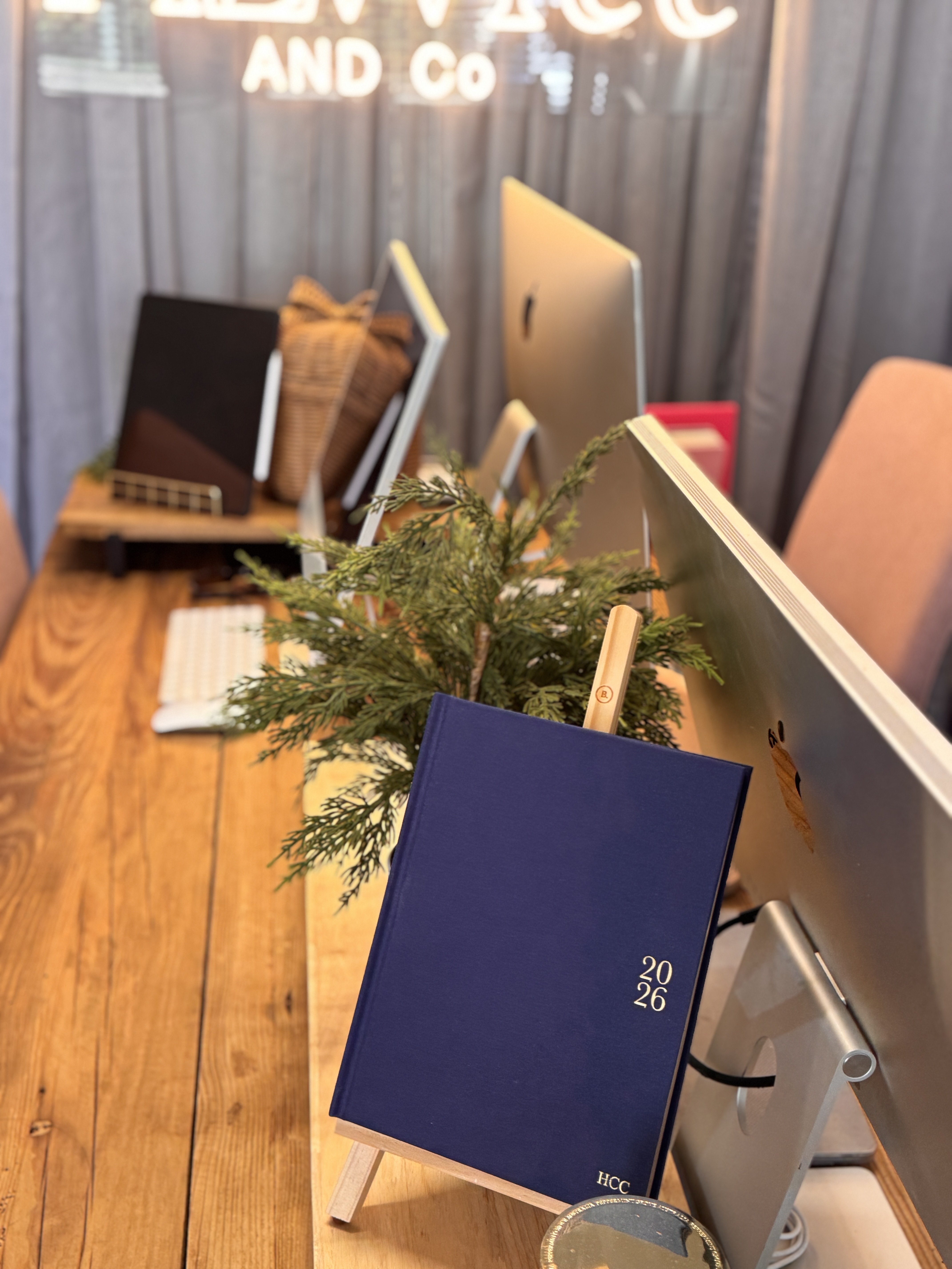 Blue 2026 planner standing on a wooden easel on a desk with a plant and computer monitors in the background.