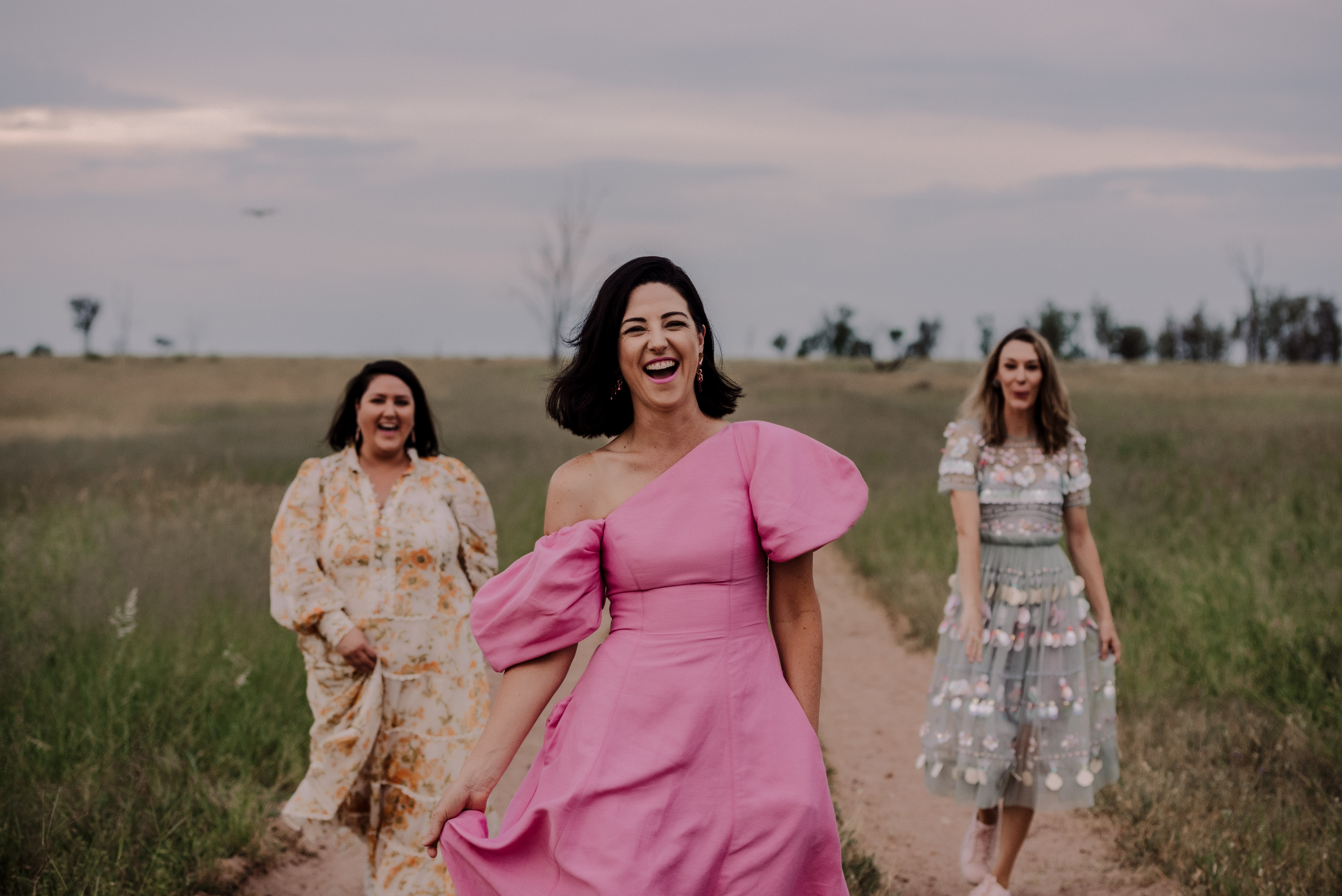 Three women walking on a dirt path in a grassy field, smiling and dressed in colorful dresses, with a cloudy sky background.