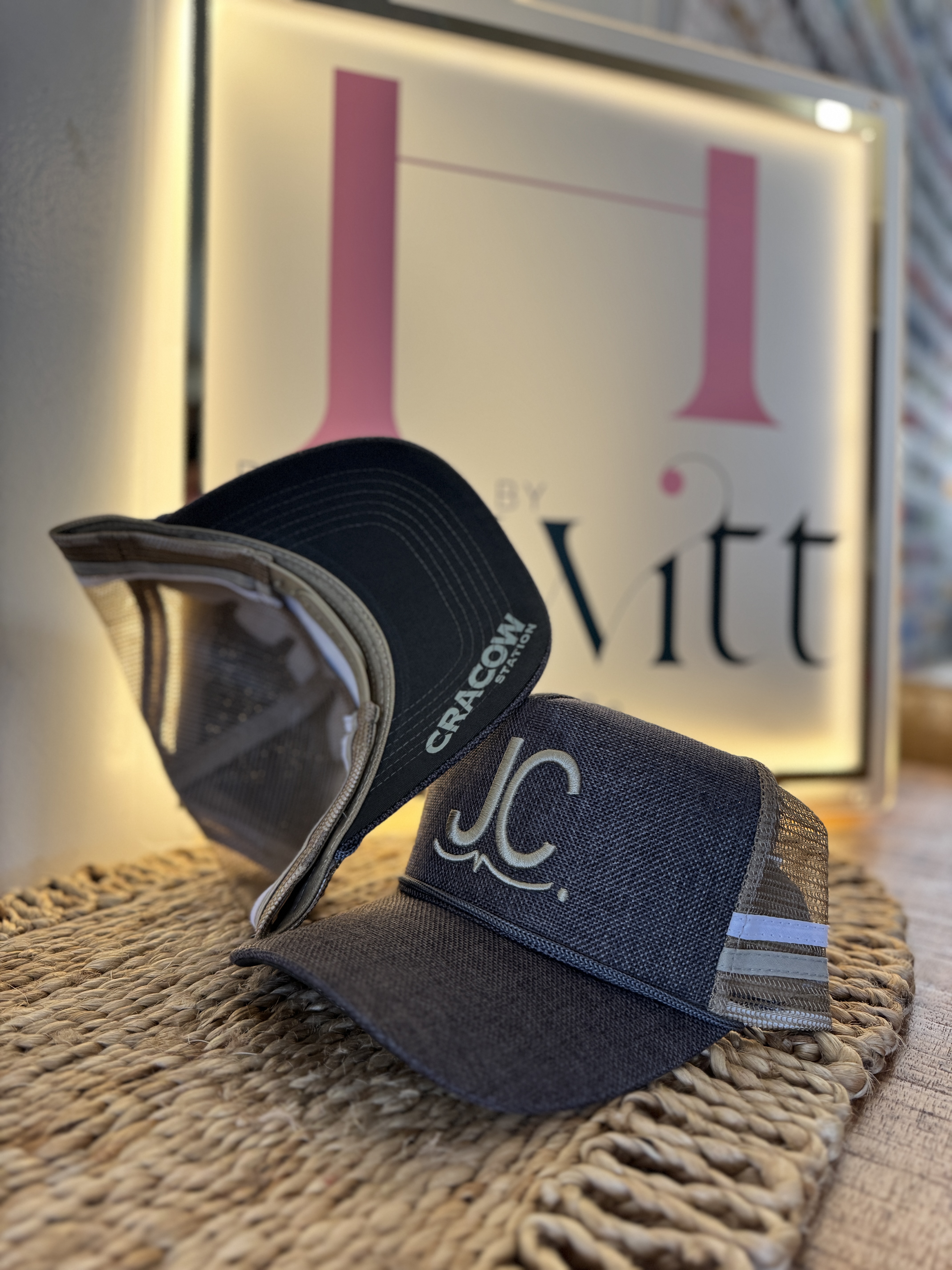 Two mesh-back baseball caps resting on a woven surface; one cap has a front logo with the initials 'J.C.' and the other shows the text 'CRACOW STATION' on the underside of its brim.