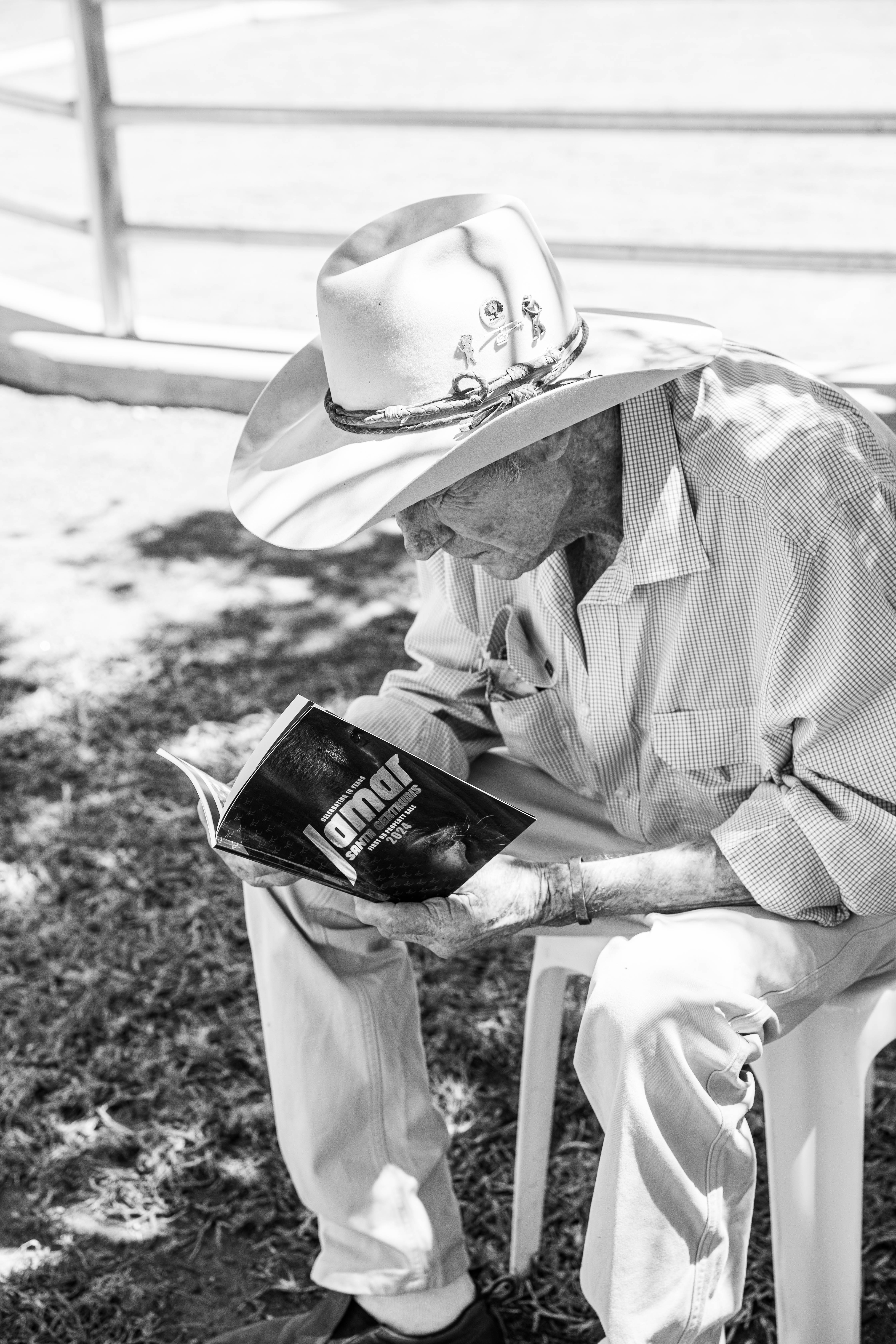 Elderly man in a wide-brimmed hat and checked shirt sitting outdoors on a plastic chair, reading a booklet titled 'Janmar'.