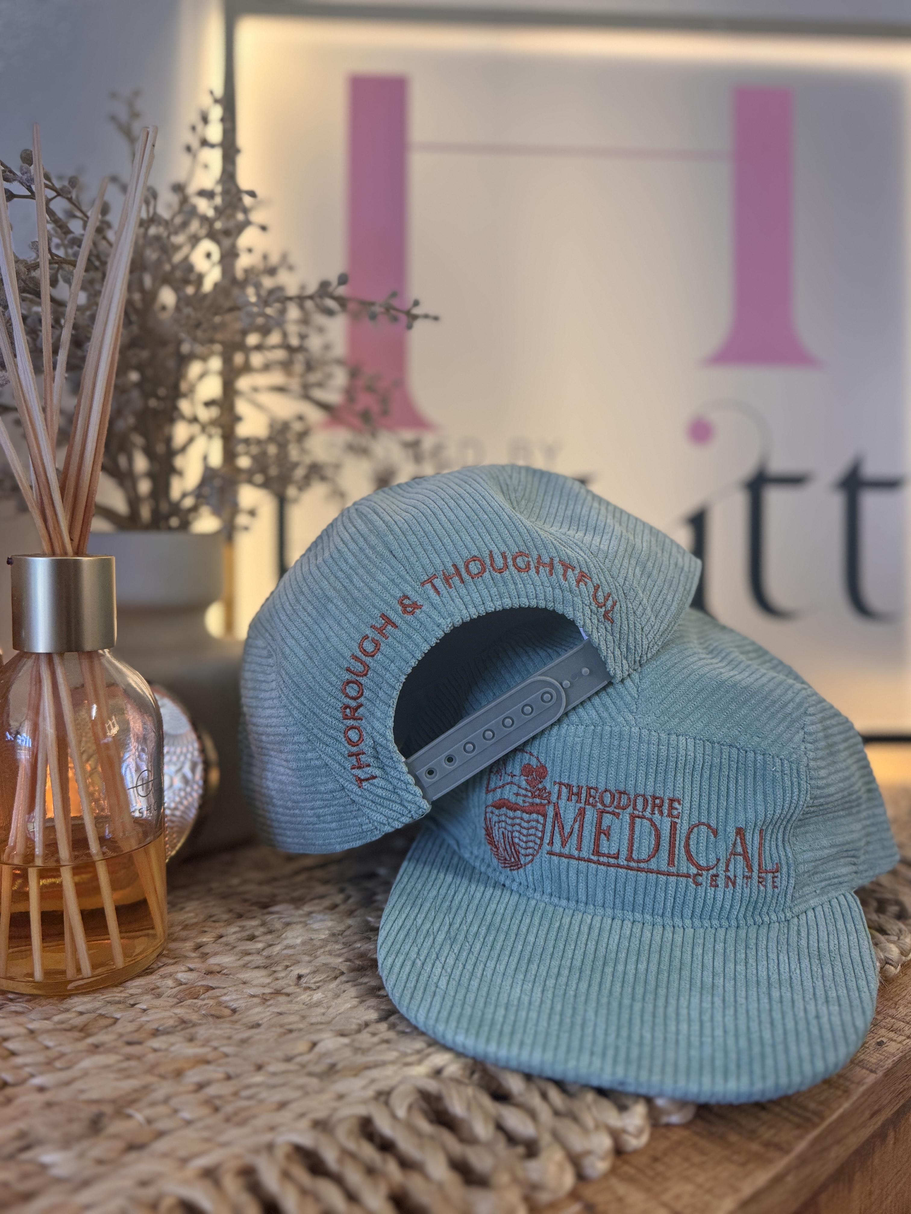 Two light blue corduroy caps stacked, one showing 'THOROUGH & THOUGHTFUL' text and the other 'THEODORE MEDICAL CENTRE' embroidery on a woven surface, with a diffuser and dried plants in the background.