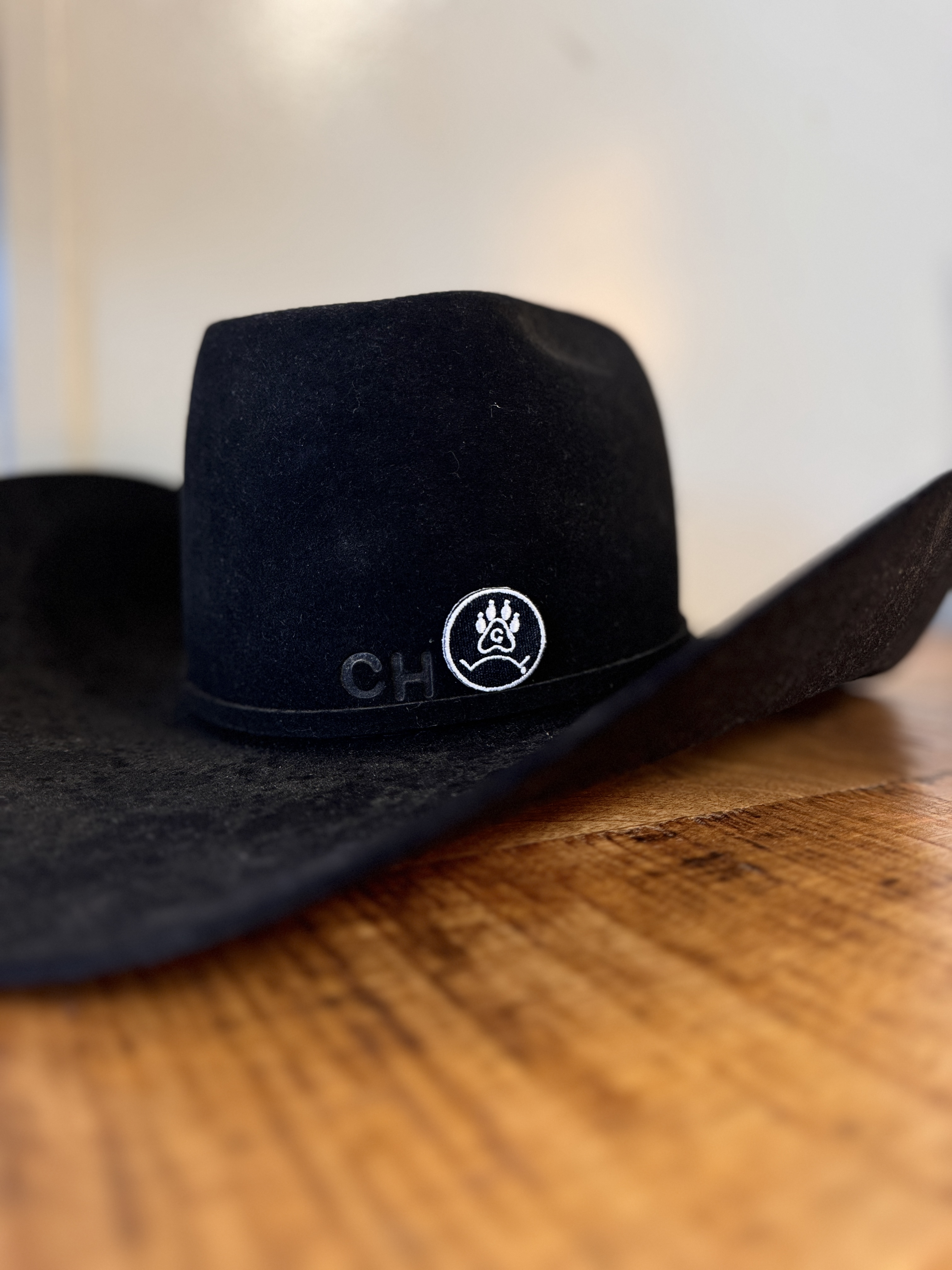 Black cowboy hat with a white embroidered paw print logo resting on a wooden surface.