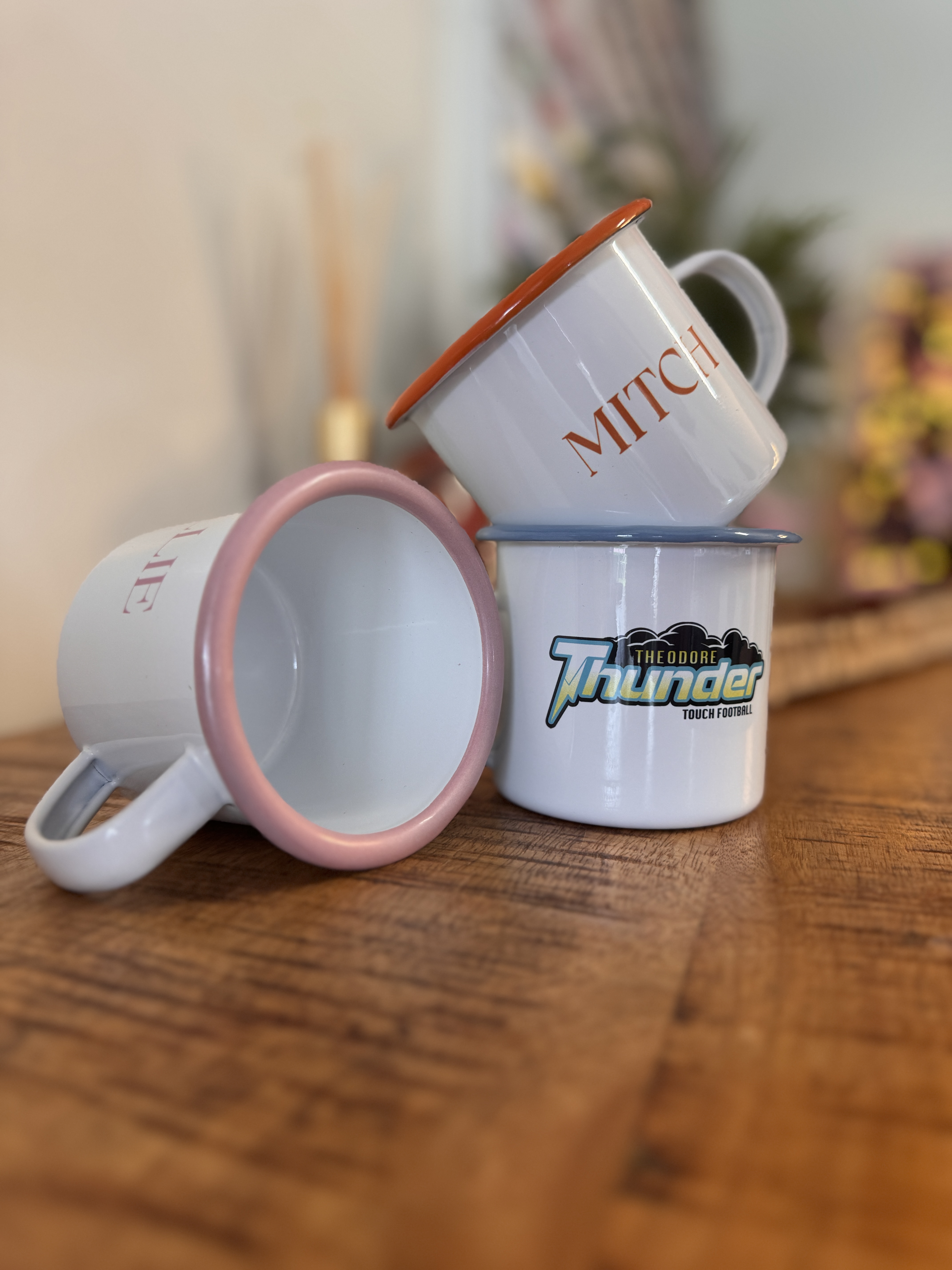 Three white enamel mugs with colored rims stacked on a wooden table, featuring text including 'MITCH' and 'THEODORE Thunder Touch Football'.