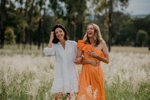 Two women laughing and walking arm in arm in a grassy field, one in a white dress and the other in an orange one-shoulder dress.