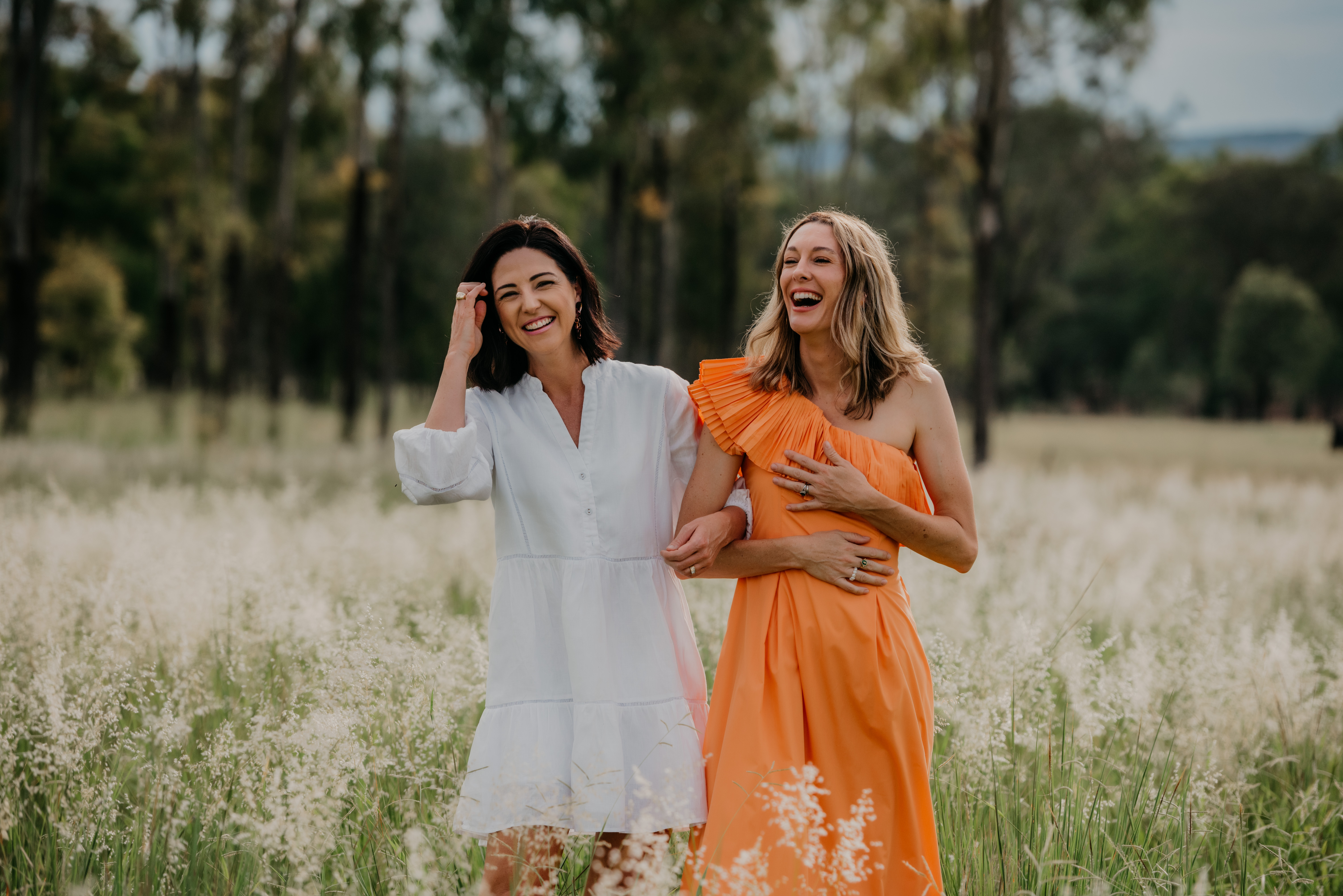 Two women laughing and walking arm in arm in a grassy field, one in a white dress and the other in an orange one-shoulder dress.