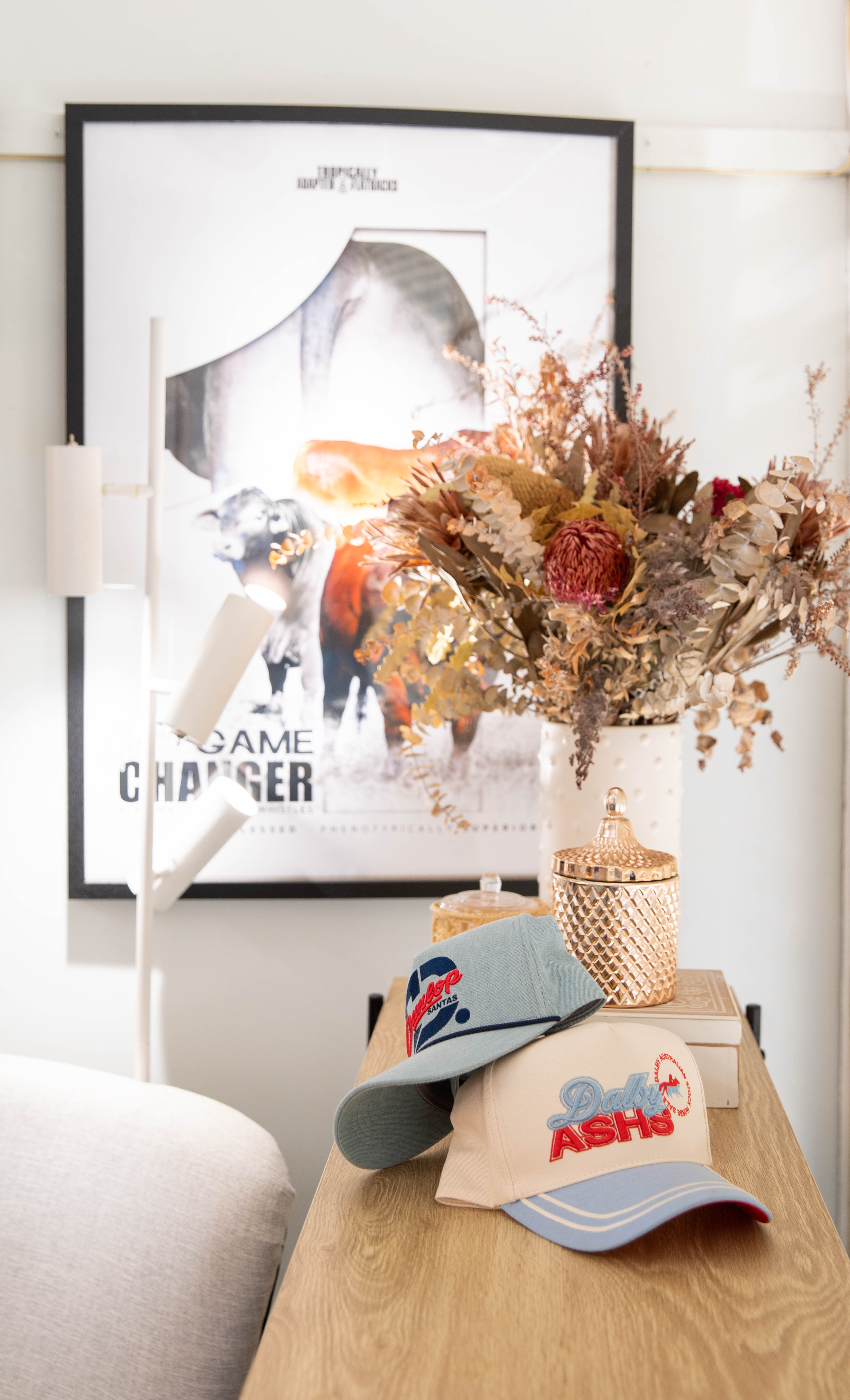 Wooden table with two baseball caps, decorative jars, and a vase of dried flowers, with a framed 'Game Changer' poster and white floor lamp in the background.