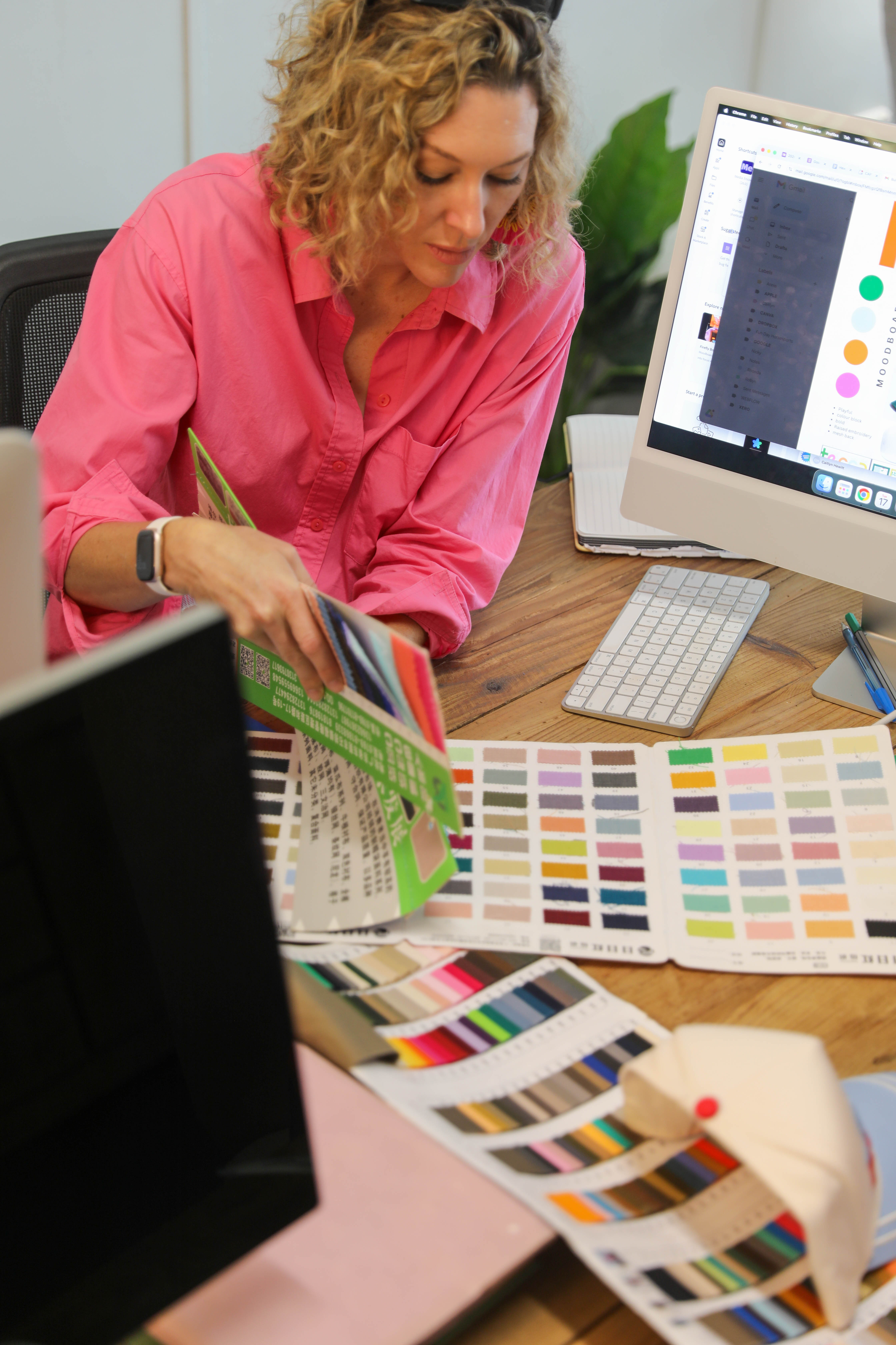 Woman in a pink shirt reviewing fabric color swatches at a wooden desk with a computer and keyboard.