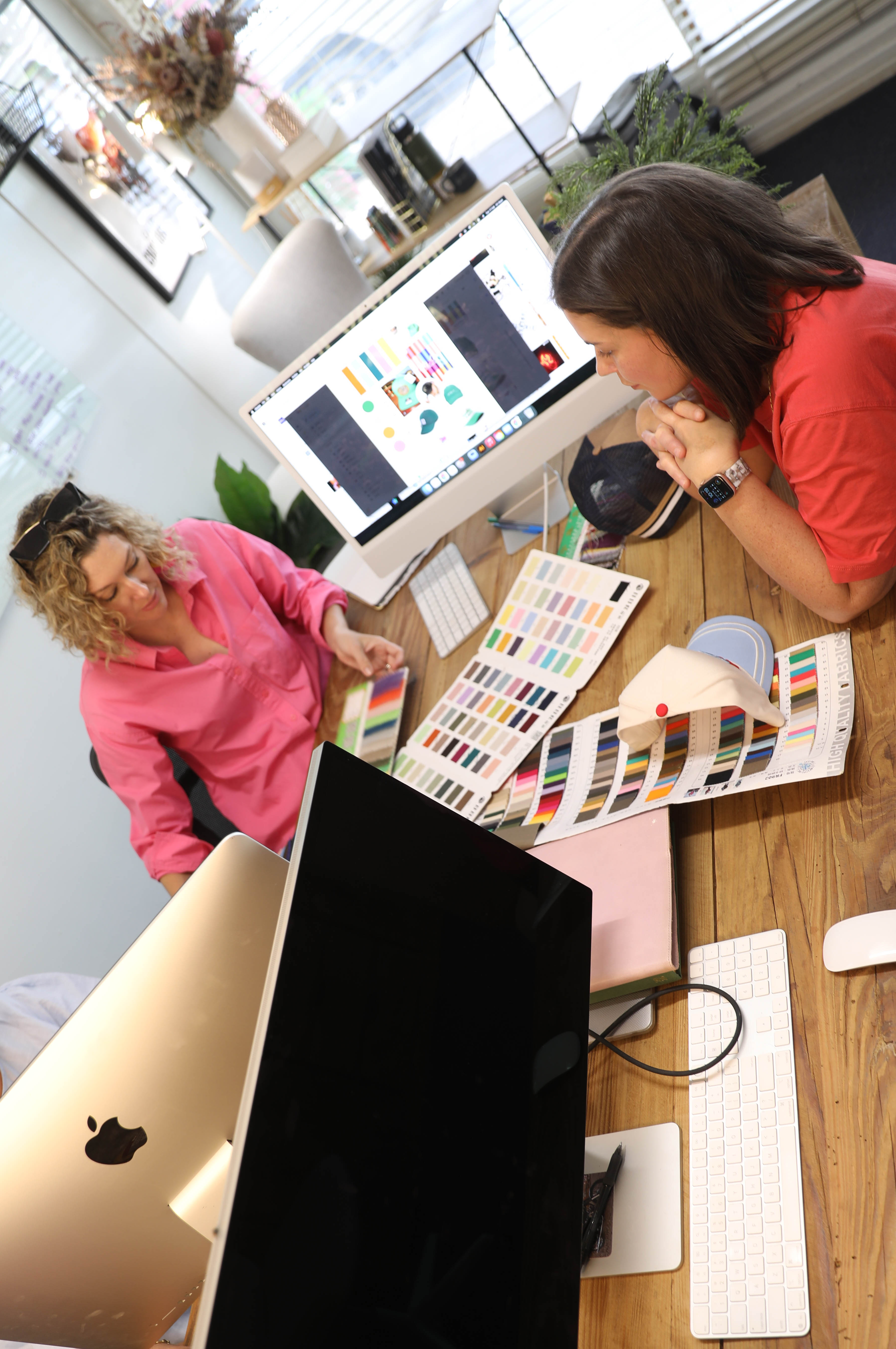 Two women at a desk reviewing fabric swatches and color samples with computer screens and design materials nearby.