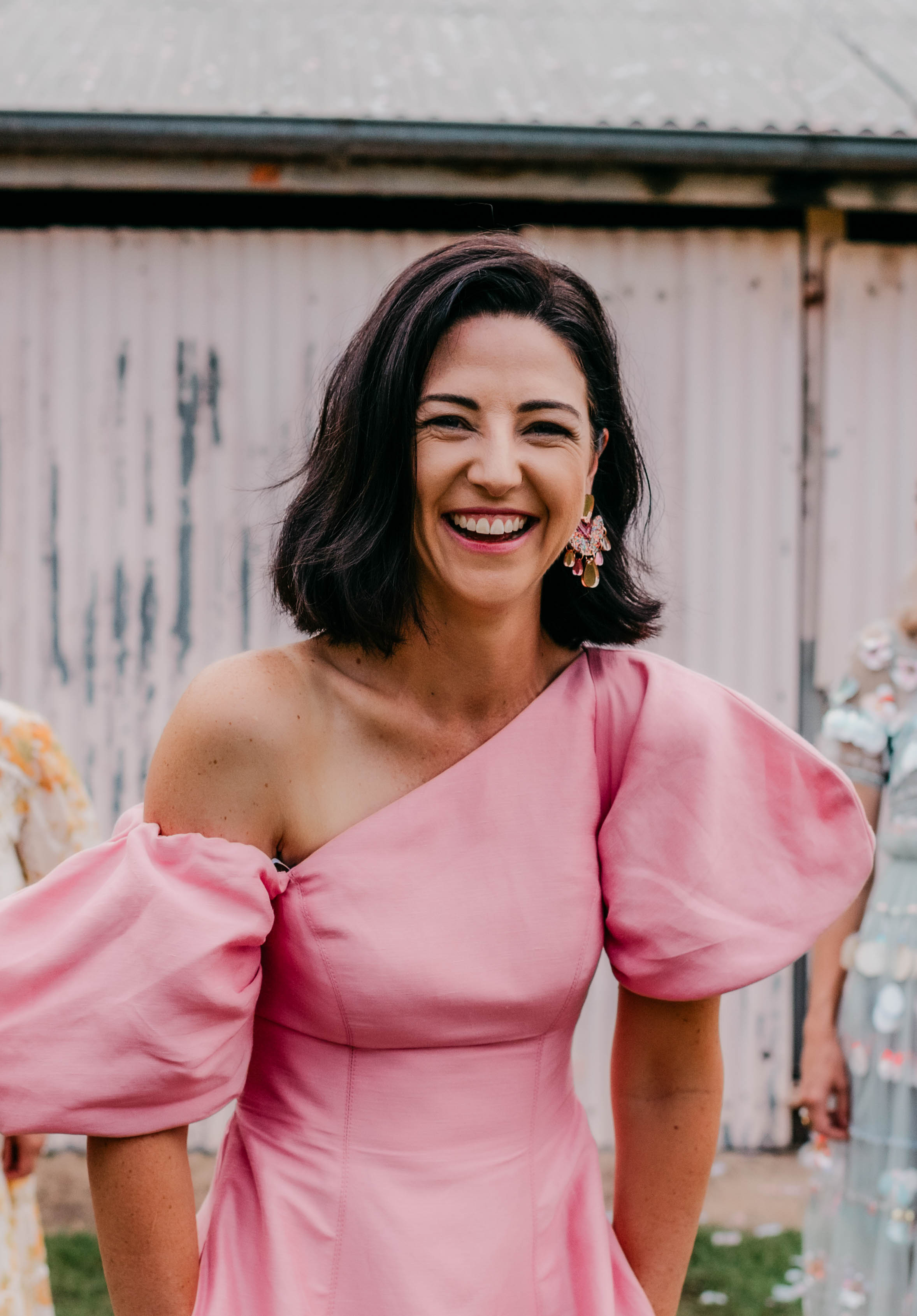 Smiling woman with dark hair wearing a pink one-shoulder dress with puffed sleeves and decorative earrings.