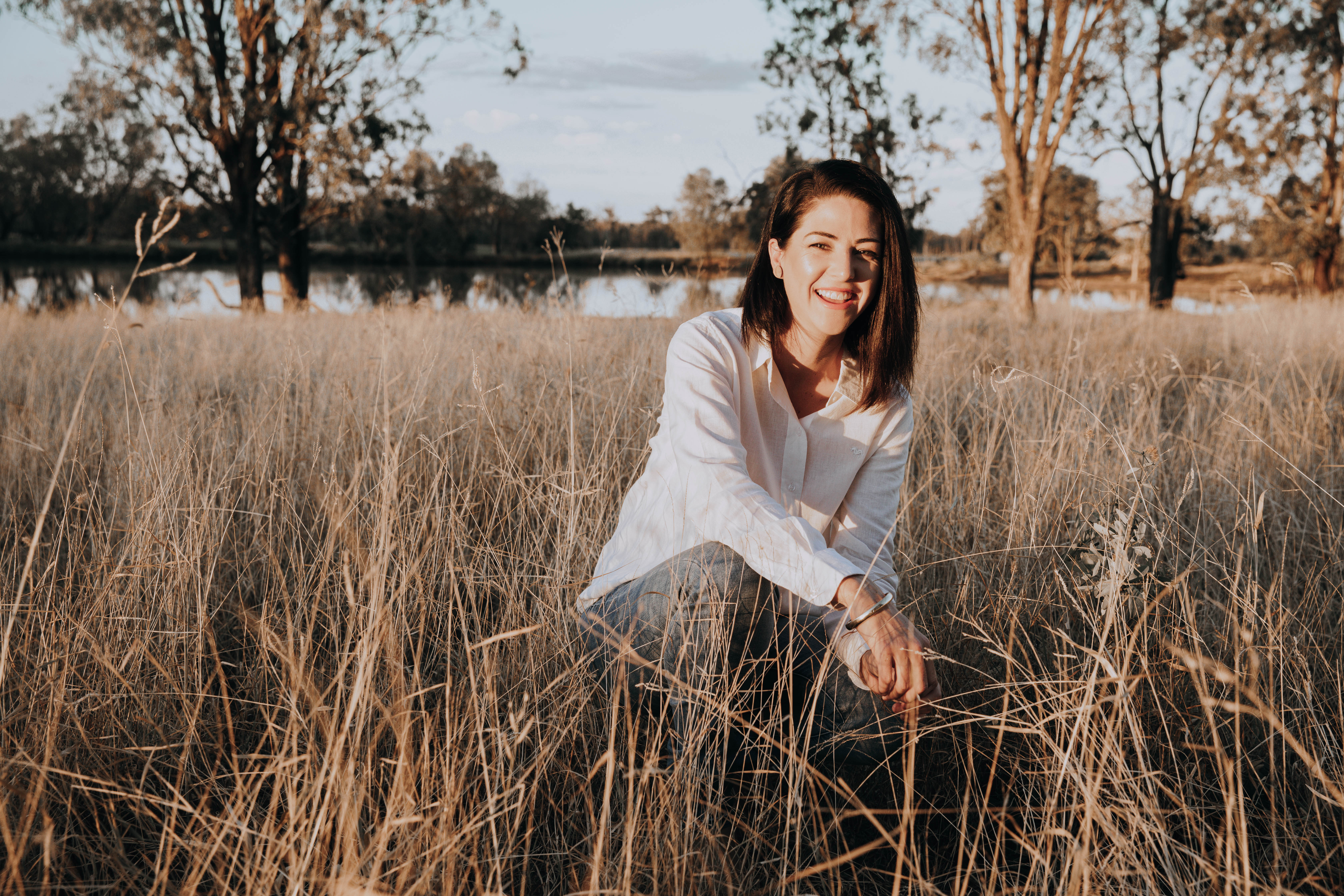Smiling woman with dark hair in a white shirt squatting in tall dry grass by a lake with trees in the background.