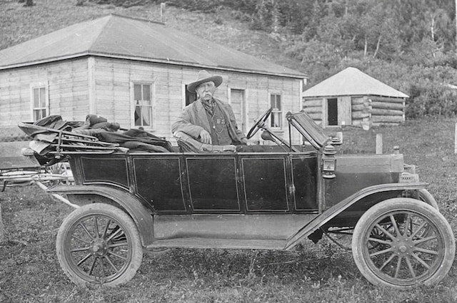 Old man standing behind his old model T looking car, with a log cabin in the background. Photo credit: Galt Museum & Archives