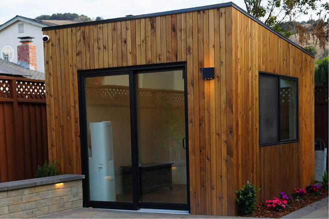 Angled view of the Minima backyard office shed by Rivolty with natural cedar siding, insulated construction, large sliding glass door, and side window.