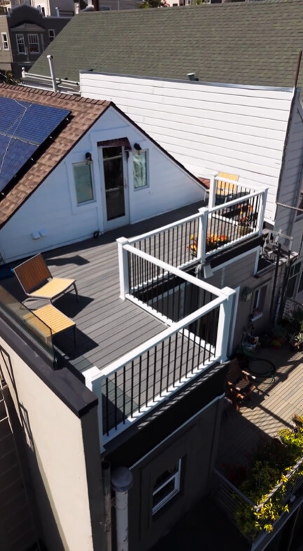 Aerial view of a modern rooftop deck with grey composite flooring and white metal railings, featuring lounge chairs and a staircase, situated next to a roof with installed solar panels.