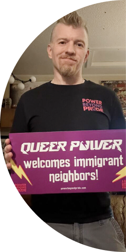 Person holding a purple sign that says 'QUEER POWER welcomes immigrant neighbors!' with lightning bolt graphics.
