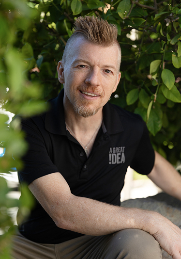 Man with a mohawk hairstyle and goatee wearing a black polo shirt with text 'A GREAT IDEA' seated outdoors among green foliage.