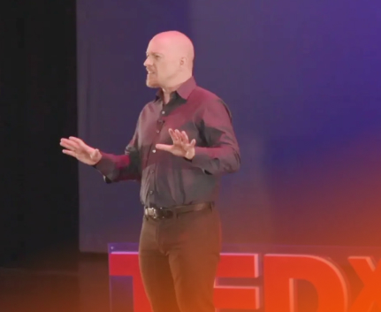 Bald man in dark shirt speaking on stage with TEDx logo partially visible in front.