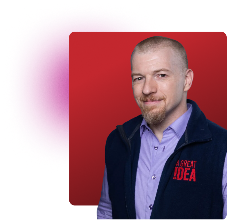 Smiling man with a short beard and mustache, wearing a light purple shirt and a dark vest with 'A GREAT IDEA' embroidered in red, against a red geometric background.