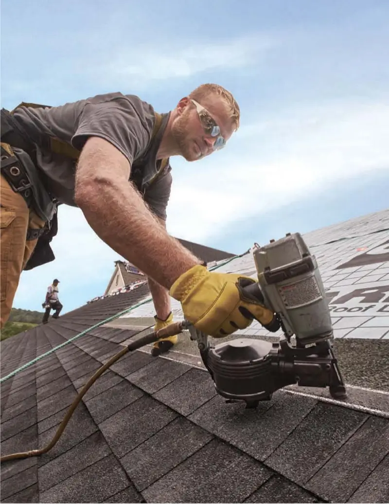 Roofer wearing yellow gloves and safety glasses using a nail gun to install shingles on a roof under a clear sky.