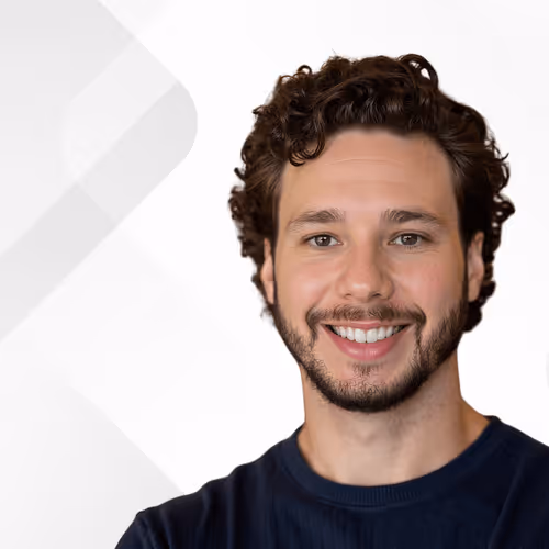 Smiling young man with curly brown hair and beard wearing a dark blue shirt against a white background.