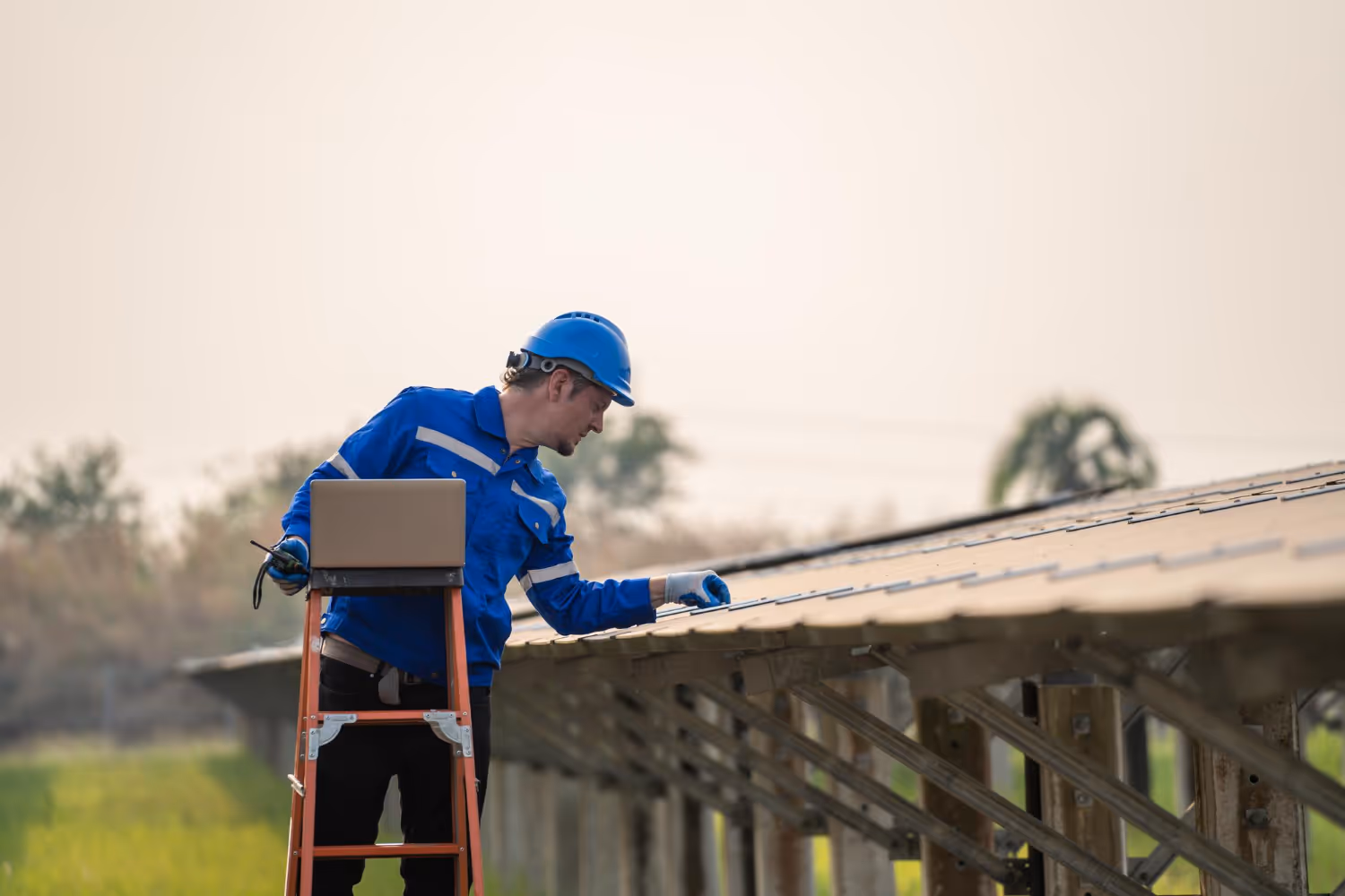 Worker in blue safety gear inspecting solar panels outdoors while standing on a ladder with a laptop.