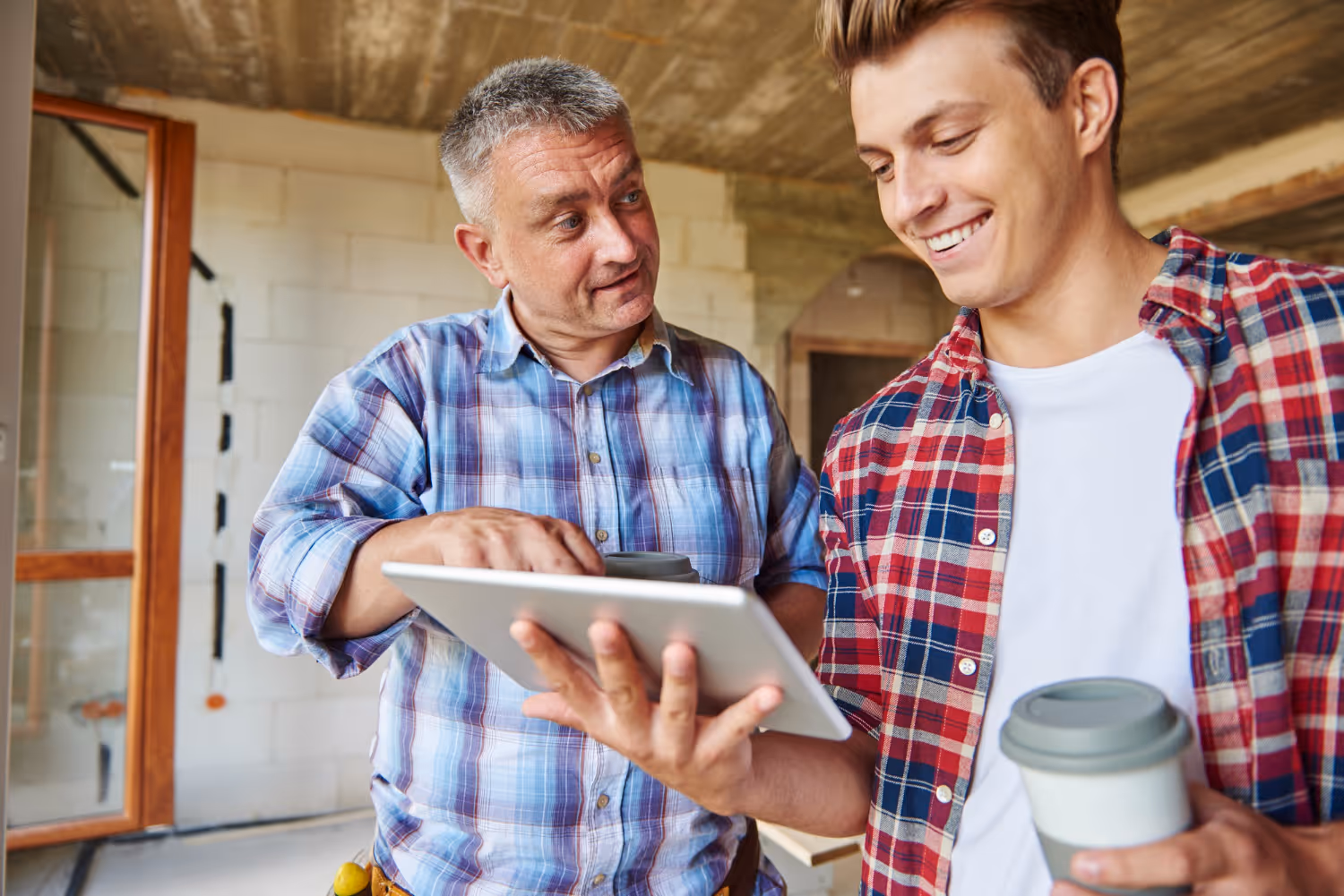 Two men in casual plaid shirts looking at a tablet, one holding a reusable coffee cup, in a partially constructed room.