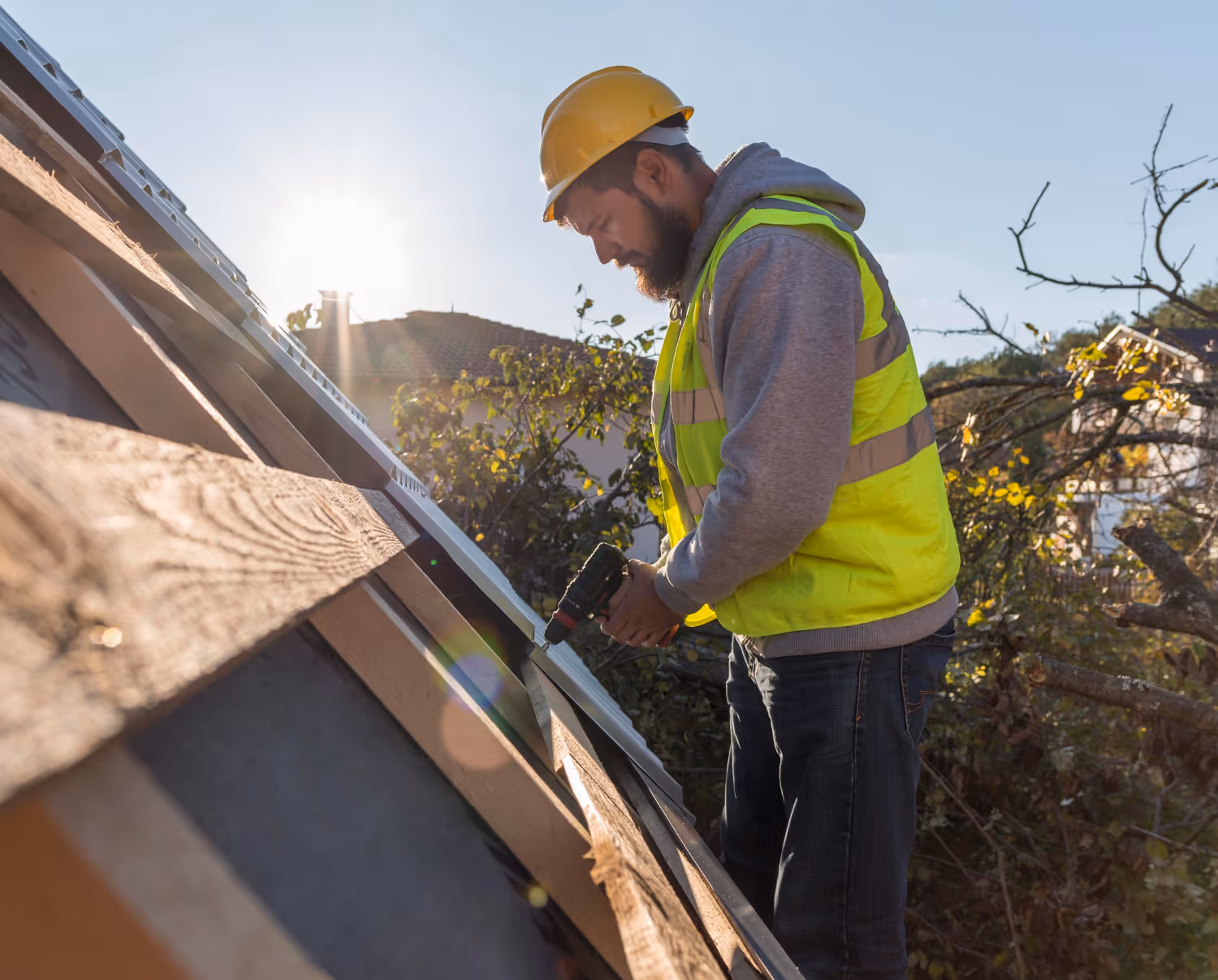 Construction worker in a yellow safety vest and hard hat installing a panel on a sloped roof using a power drill during sunny weather.