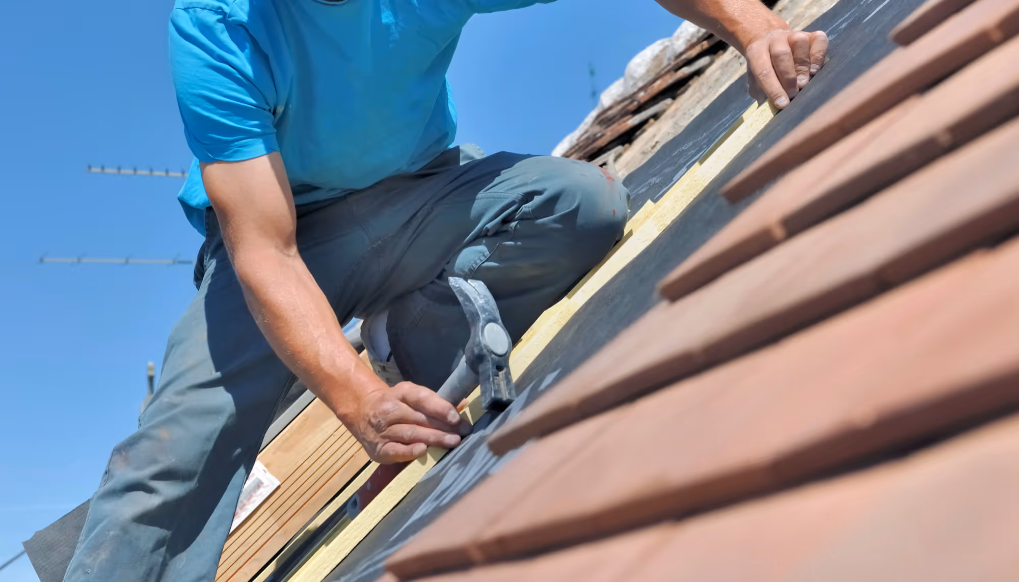 Worker in blue shirt and gray pants kneeling on a roof, holding a hammer and installing roofing tiles under a clear blue sky.