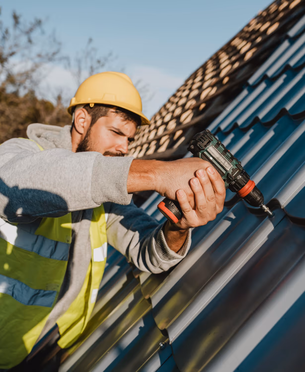Construction worker wearing a yellow hard hat and safety vest using a power drill on a metal roof.