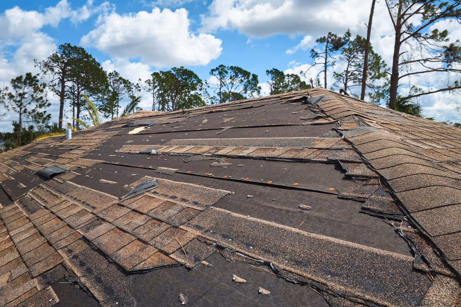 Roof with extensively damaged and missing shingles under a partly cloudy sky with trees in the background.
