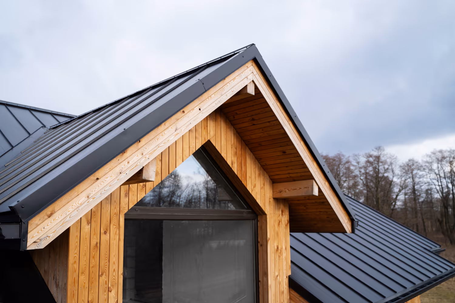 Wooden house gable with a large triangular glass window and a black metal roof under a cloudy sky.