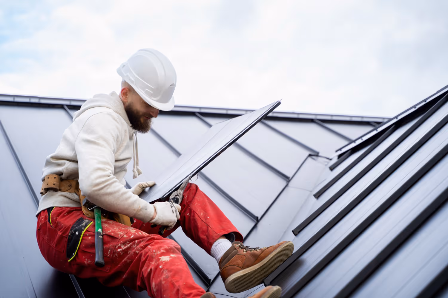 Worker in a white hard hat and red pants installing a solar panel on a metal rooftop.