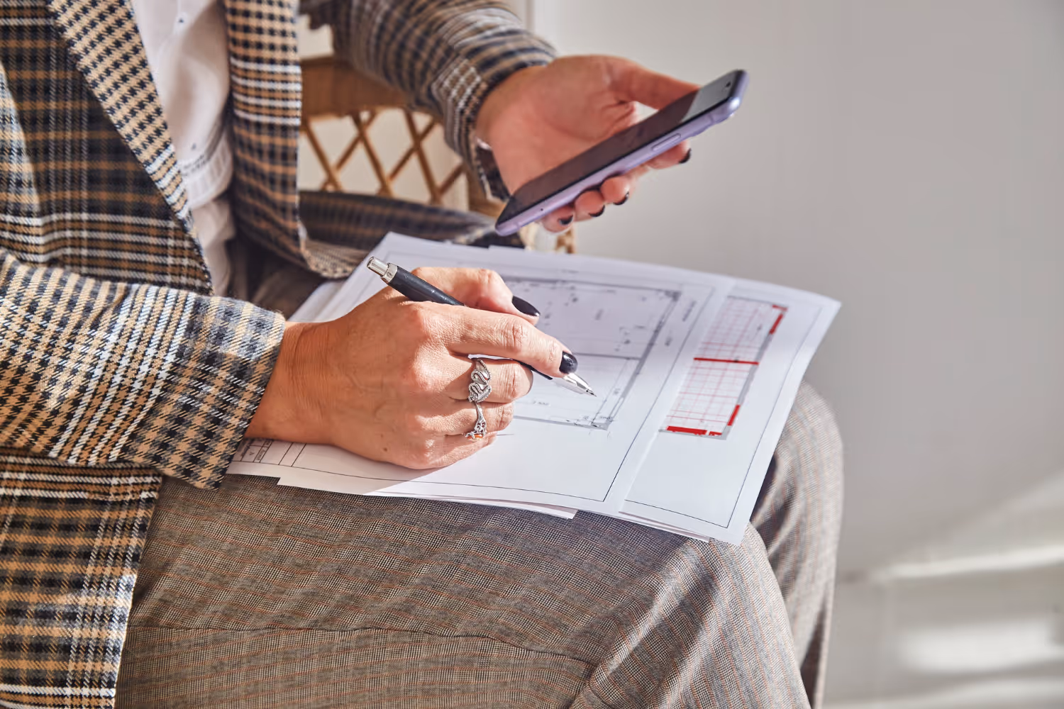 Person holding a smartphone in one hand and marking architectural blueprints with a pen in the other hand.