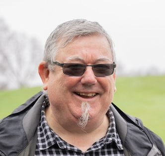 Smiling young man with short brown hair wearing a colorful patterned shirt against a muted pink background.