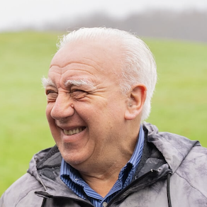 Smiling man with short dark hair and beard wearing a dark shirt against a beige background.