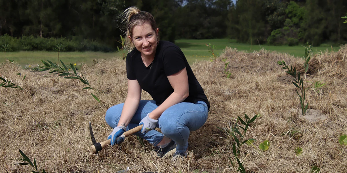 Woman kneeling outdoors planting small trees in dry soil, holding planting tool and wearing gloves.