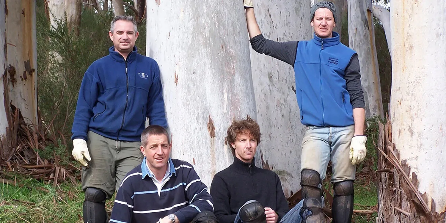 Four men wearing protective knee pads and gloves posing outdoors among large eucalyptus trees.