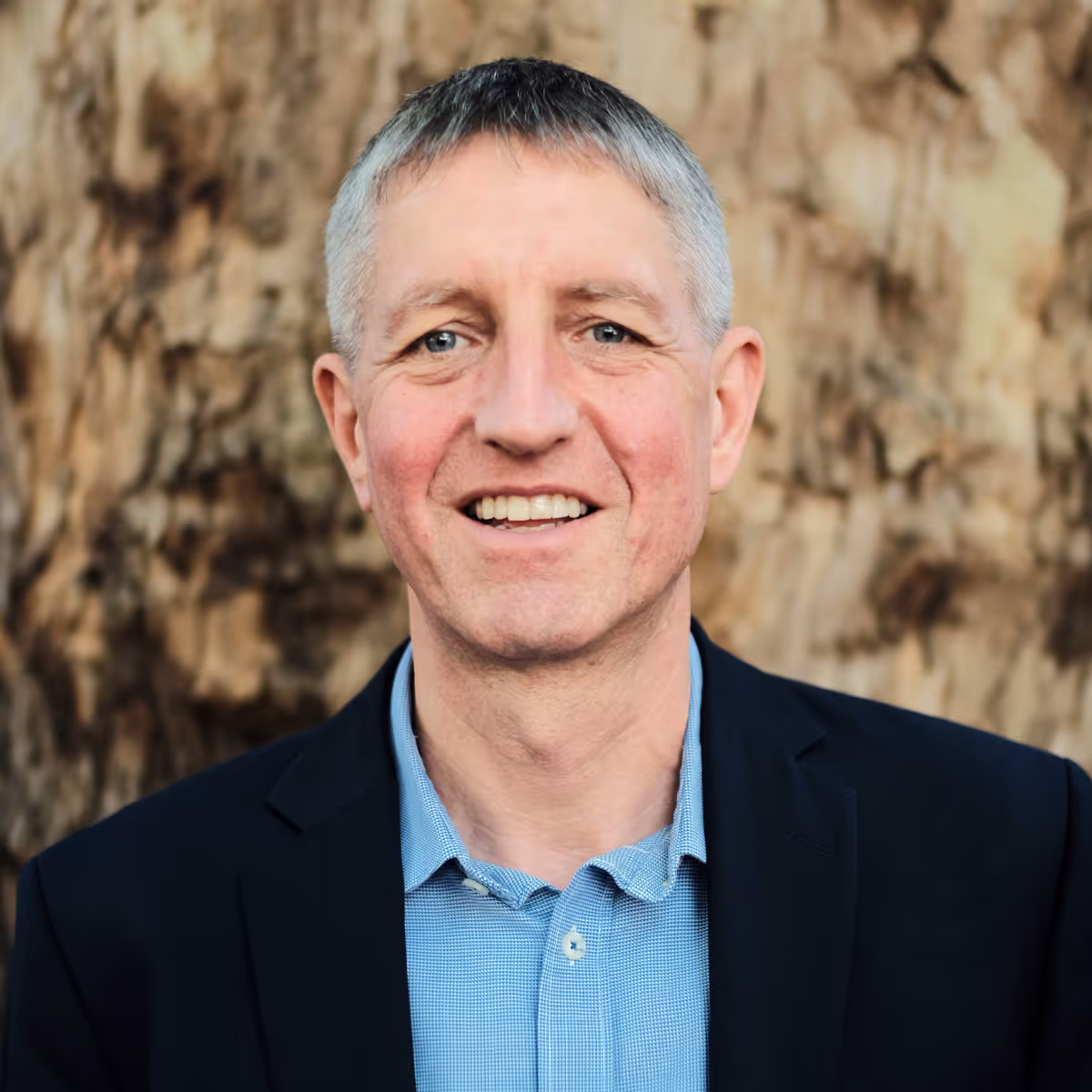 Smiling middle-aged man with short gray hair wearing a blue shirt and navy blazer against a tree background.
